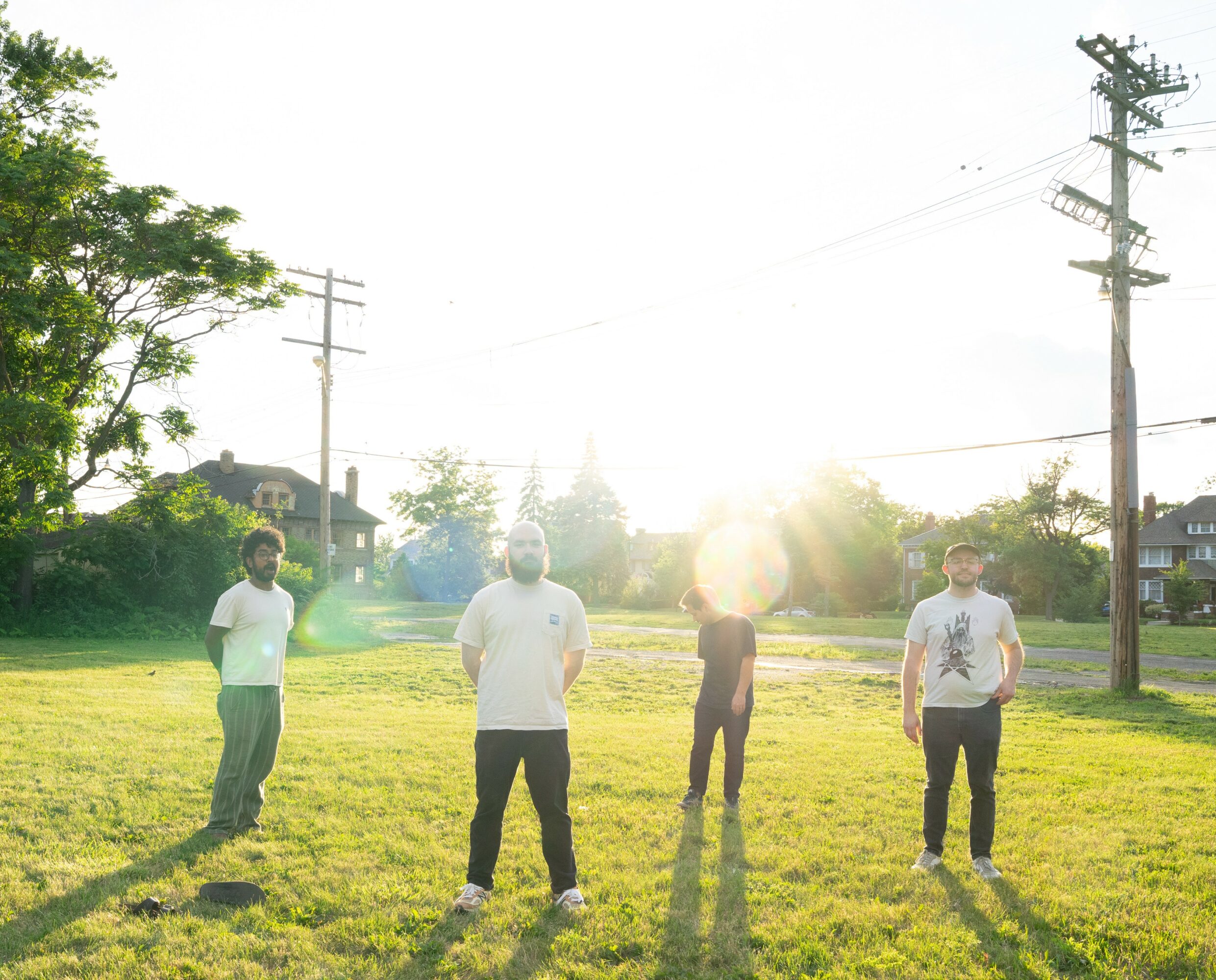 Four people standing on grass with sunlight behind them, trees, houses, and utility poles in the background.