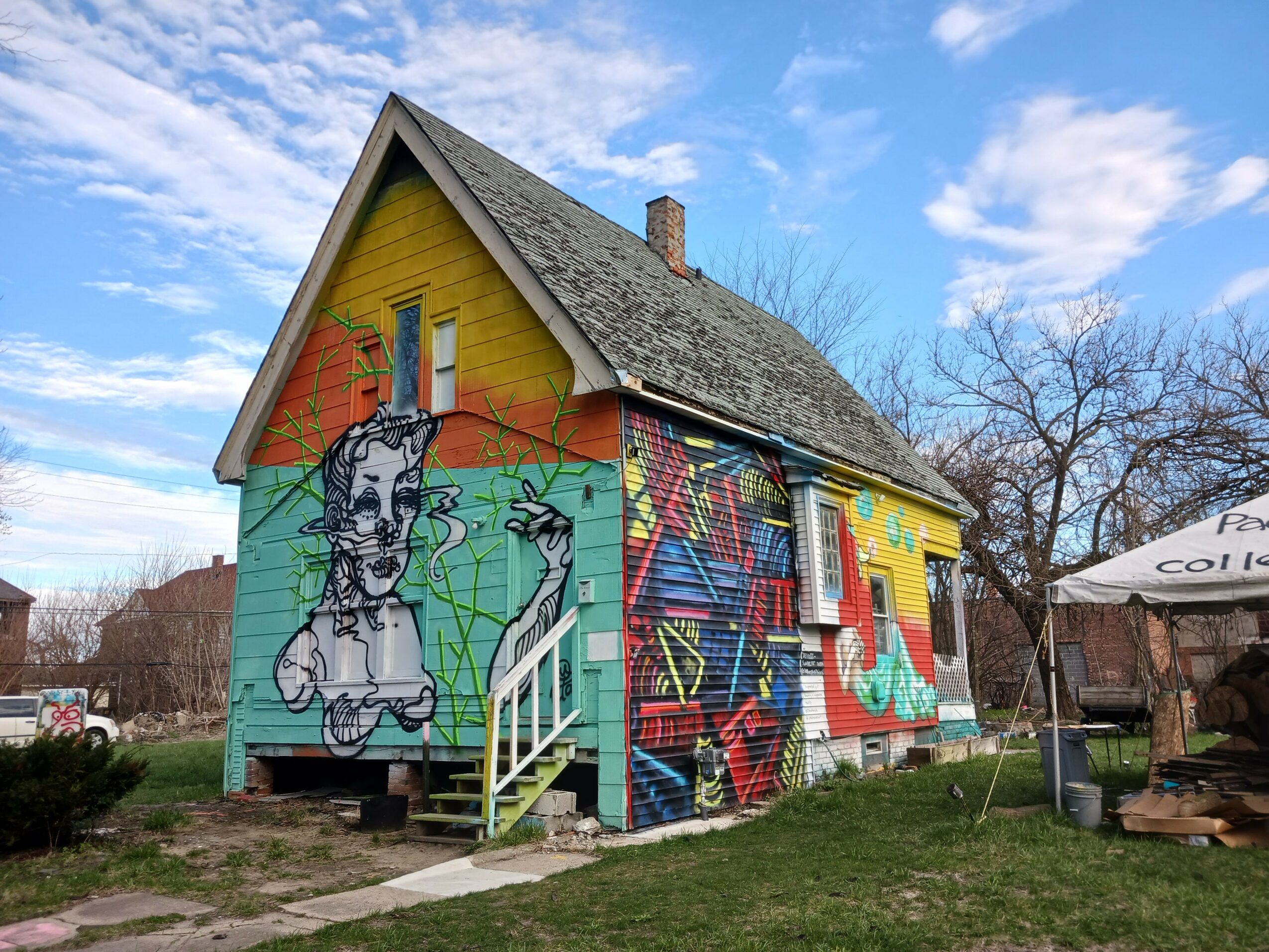 House with colorful mural, stairs, and a grassy yard, under a partly cloudy sky.