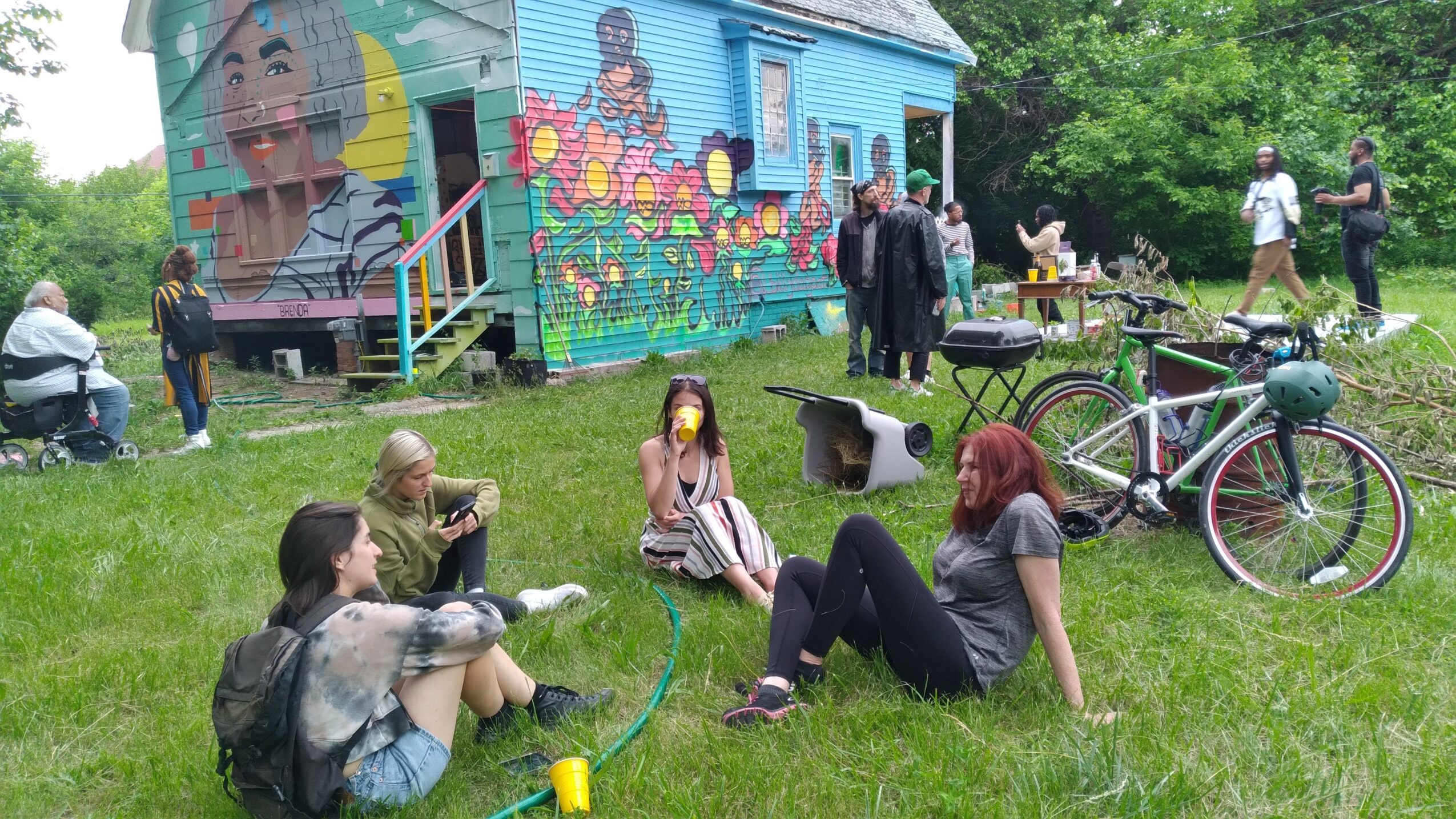 People sitting and chatting on grass near a colorful mural building, with bicycles and trees in the background.