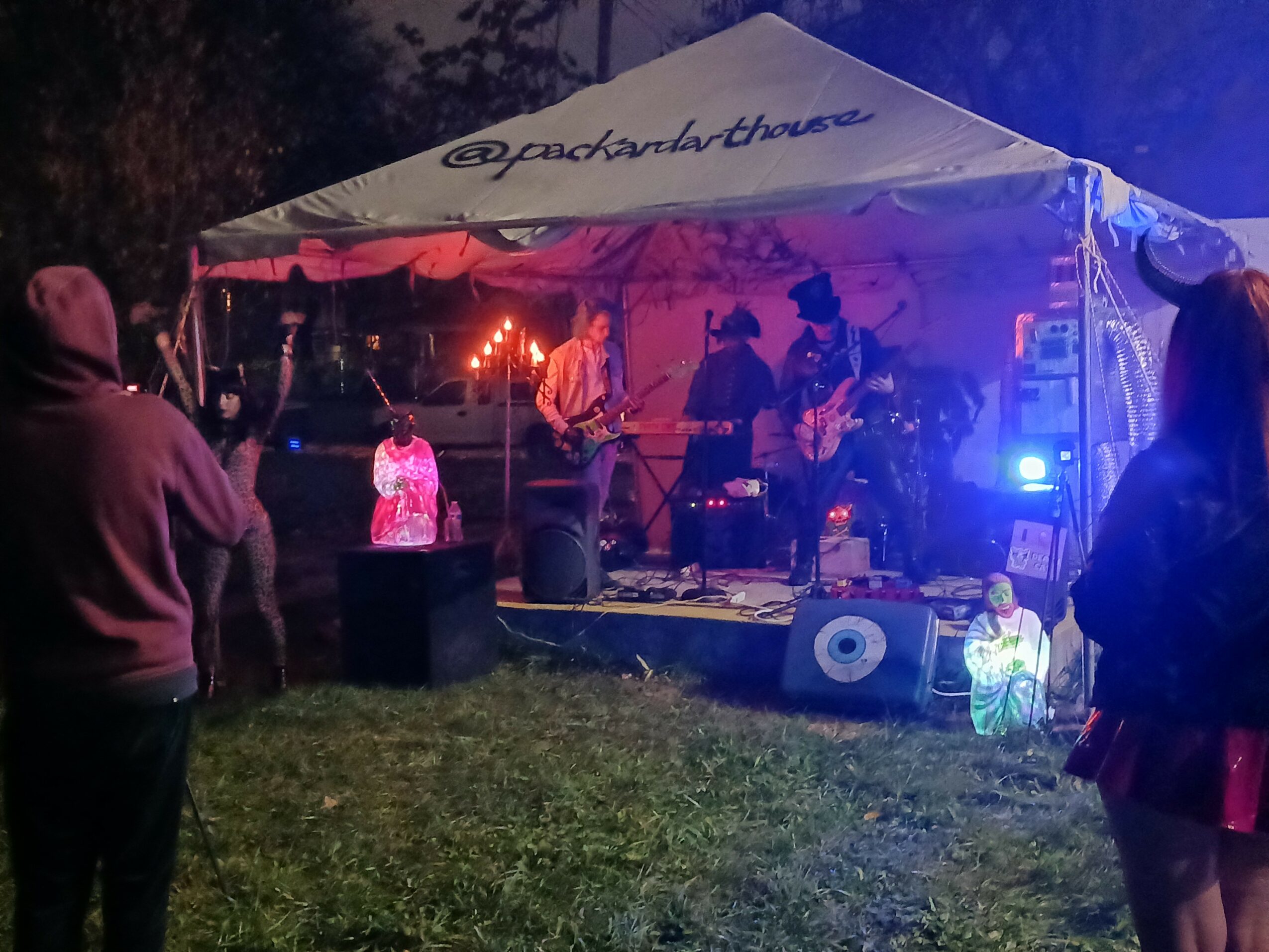 People watching a band perform on an outdoor stage at night under a tent, with colorful lighting and grass in foreground.