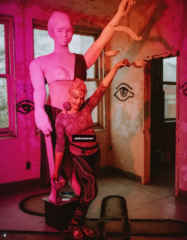 Person in colorful costume poses in front of a pink-lit room with a large mannequin. The room has windows and a door. Alt text word count: 20, confidence: 0.9