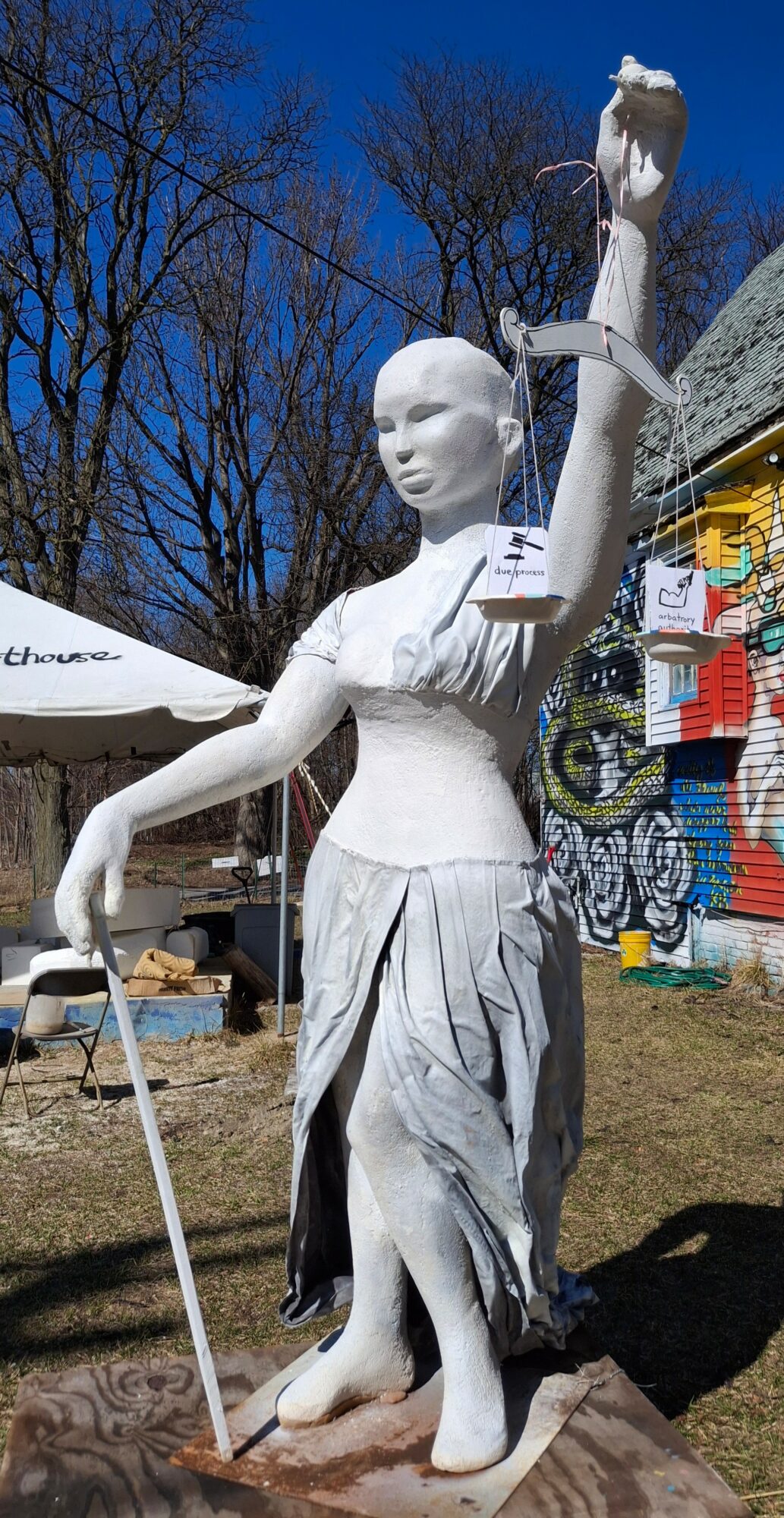 White statue of a woman holding a small object in her raised hand, standing outdoors with trees and colorful structures behind.