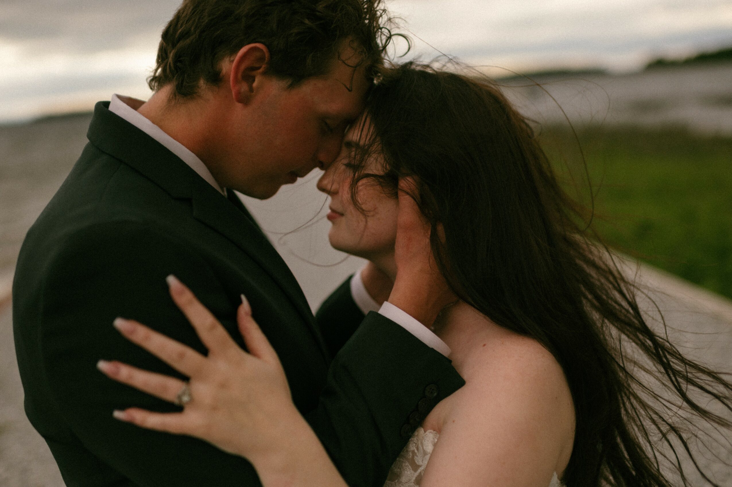 A man and woman face each other closely outdoors, with a blurred landscape background, embracing tenderly.