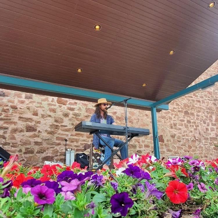 Person wearing a hat playing a keyboard outdoors with colorful flowers in foreground and brick wall background.