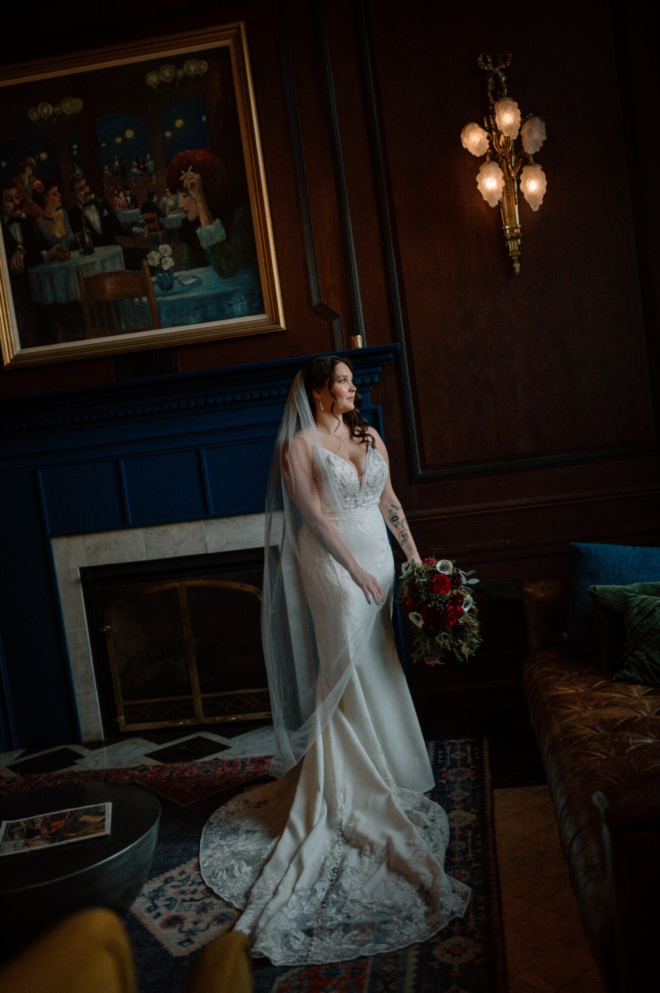 Bride in a wedding dress holding a bouquet, standing in a dimly lit room with dark wood walls and a painting above the fireplace.