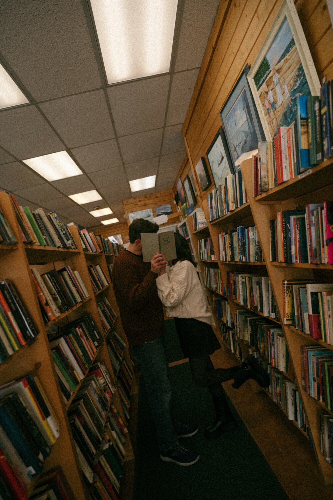 Two people browsing books in a bookstore aisle with wooden shelves and ceiling lights.