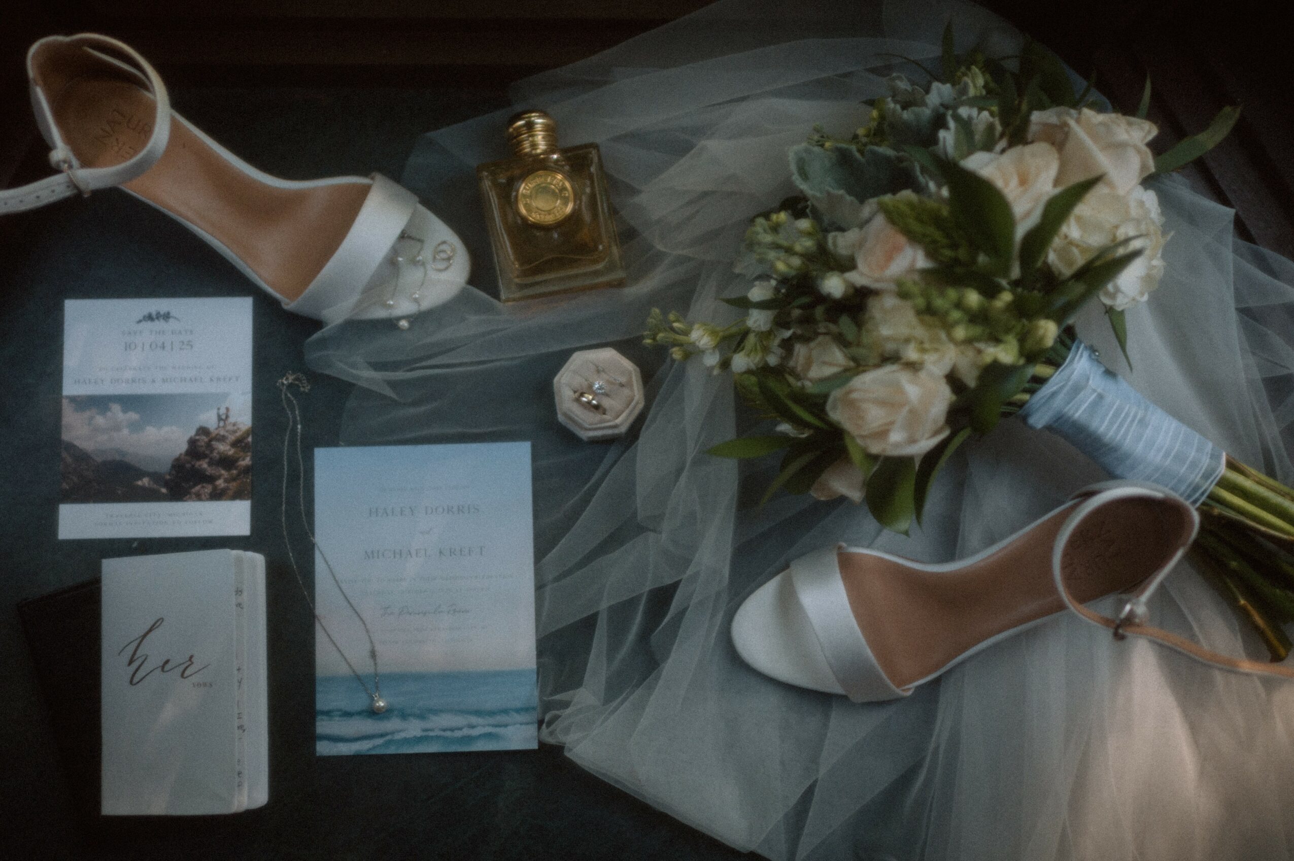 White high-heeled shoes, a bouquet of white roses, a perfume bottle, a card, and a small box on sheer fabric.