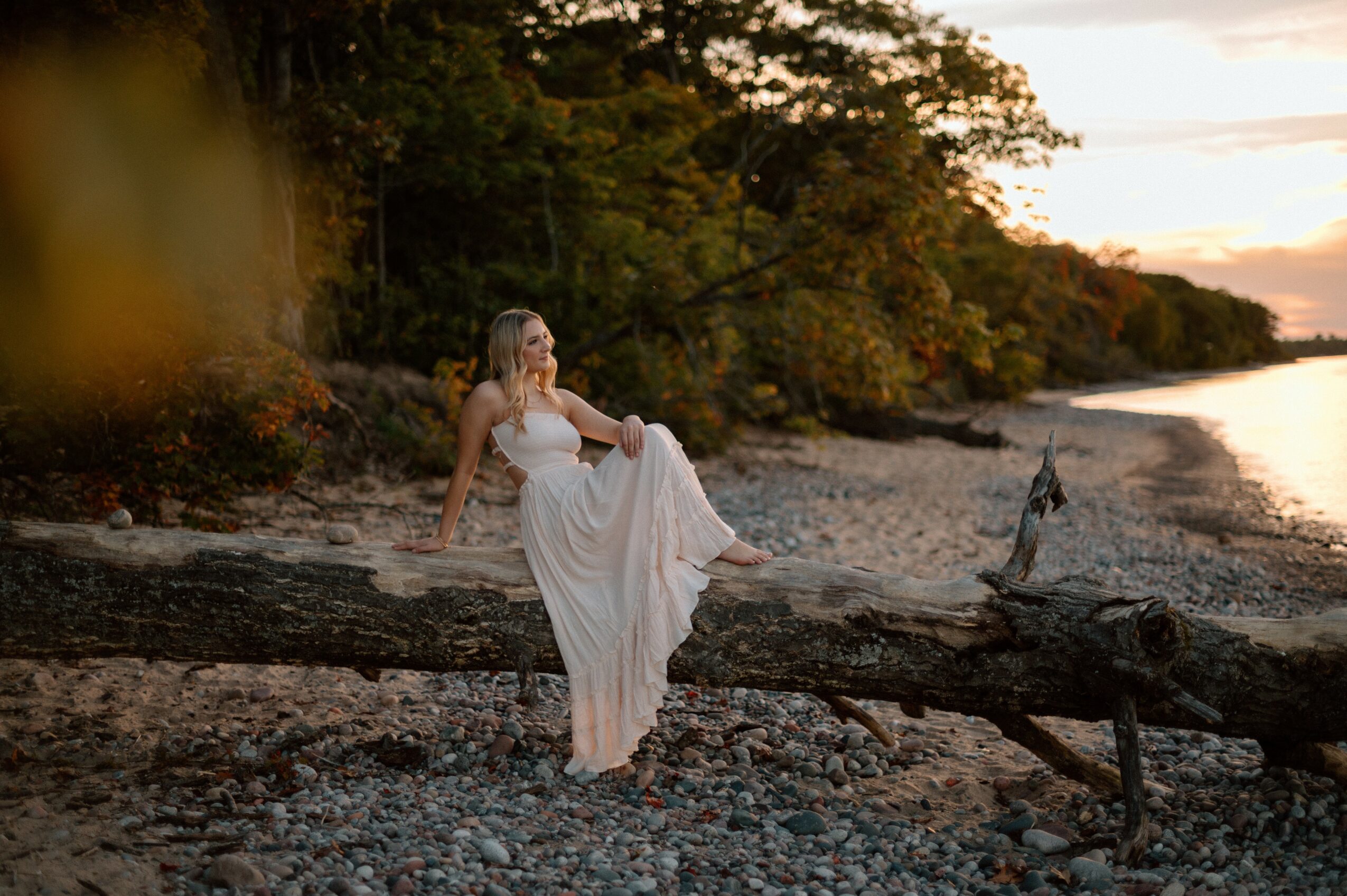Woman in a white dress sitting on a fallen tree by a river at sunset, surrounded by trees and nature.
