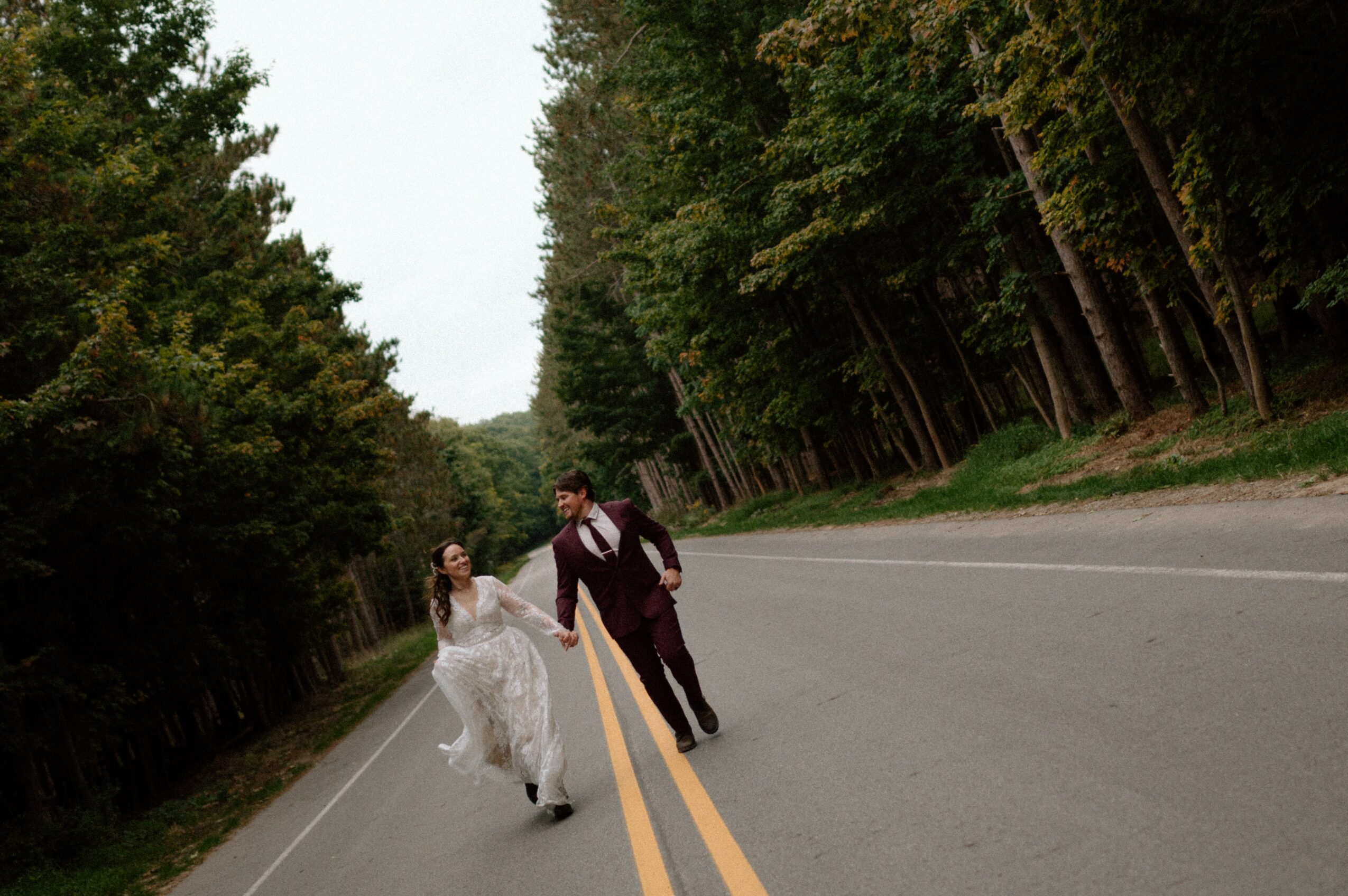 A couple walking on a two-lane road surrounded by trees, holding hands and looking at each other.