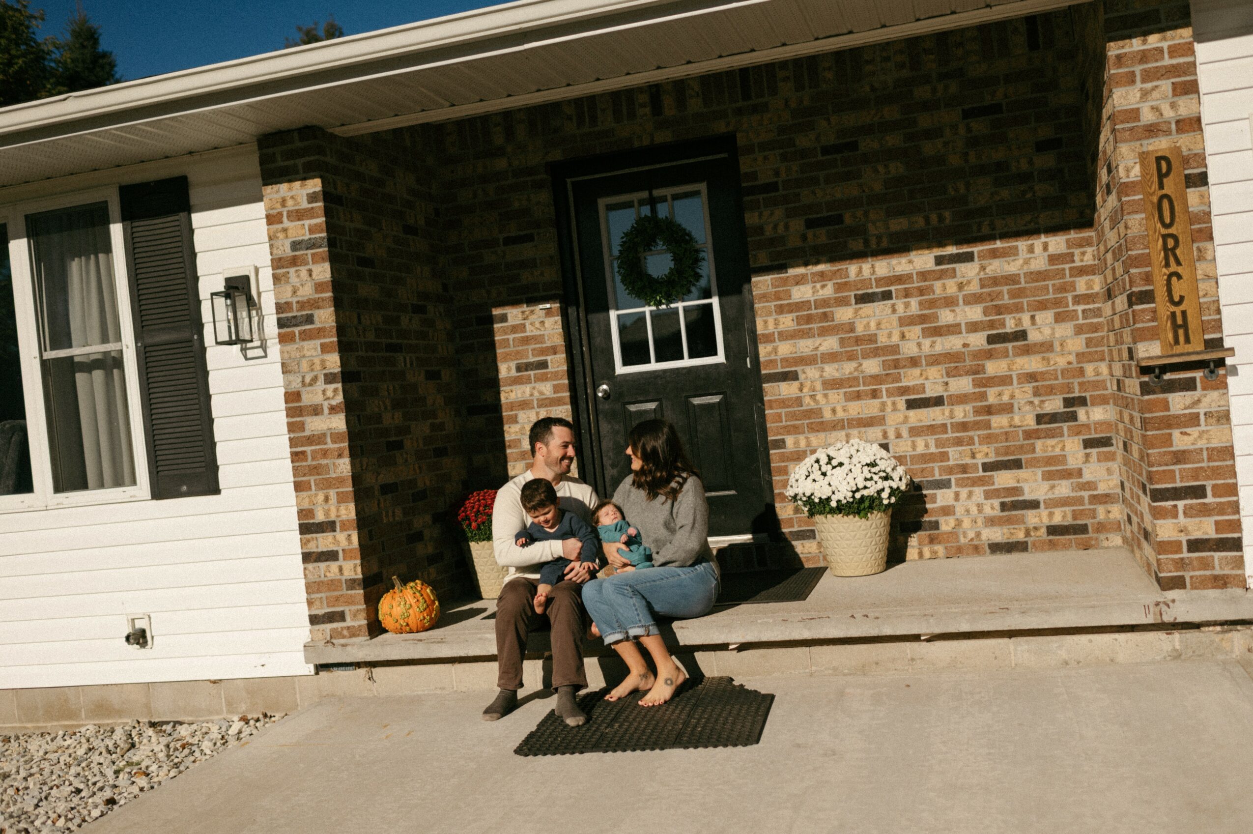 Family sitting on porch steps in front of house with brick wall, door with wreath, and potted plant, sunny day.