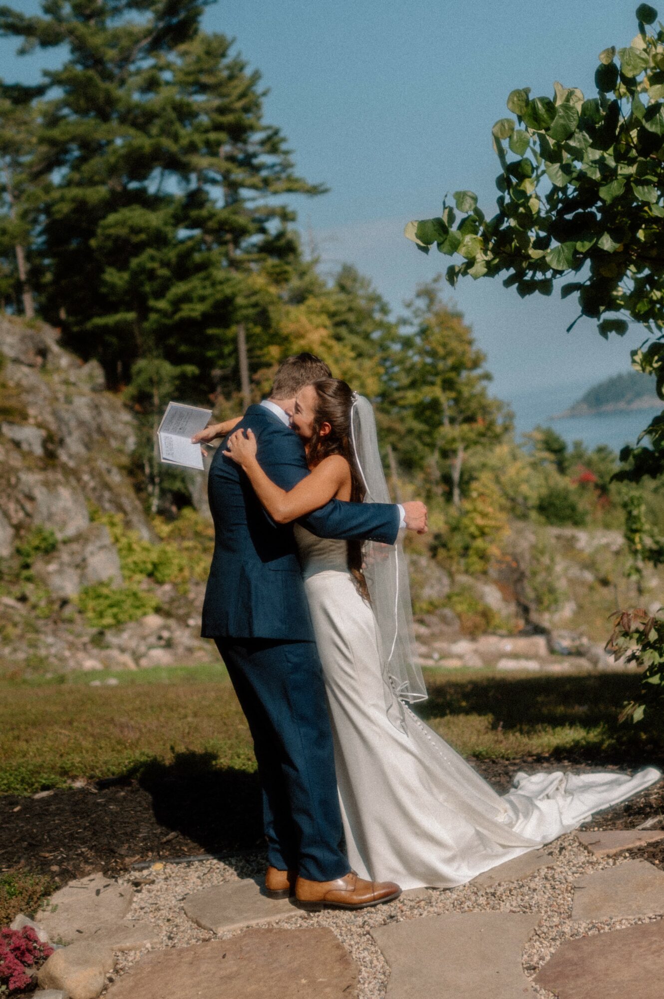 A couple in wedding attire embracing outdoors with trees and sky in background.