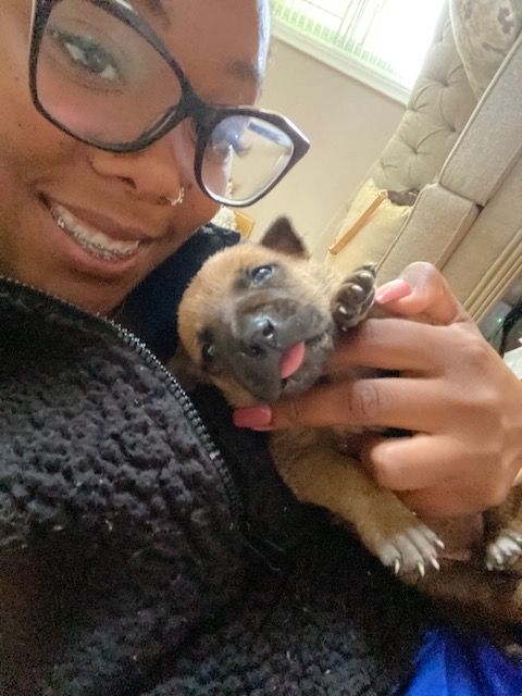 Person with glasses holding a small puppy, smiling, indoors near a window.