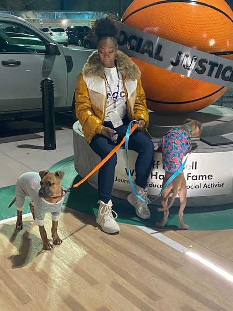 Person sitting on a bench holding a dog, with a large basketball and a woman in the background. Indoor setting.