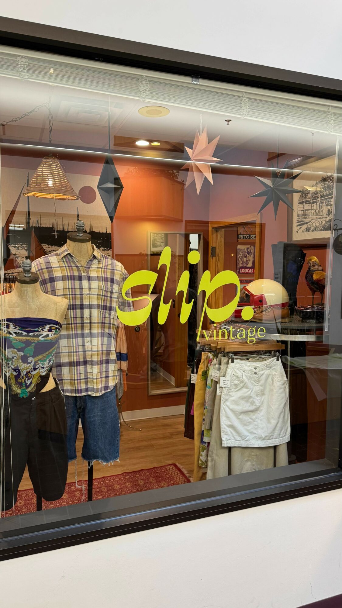Clothing store window display with mannequins wearing casual outfits, and a yellow sign reading 'slip'.