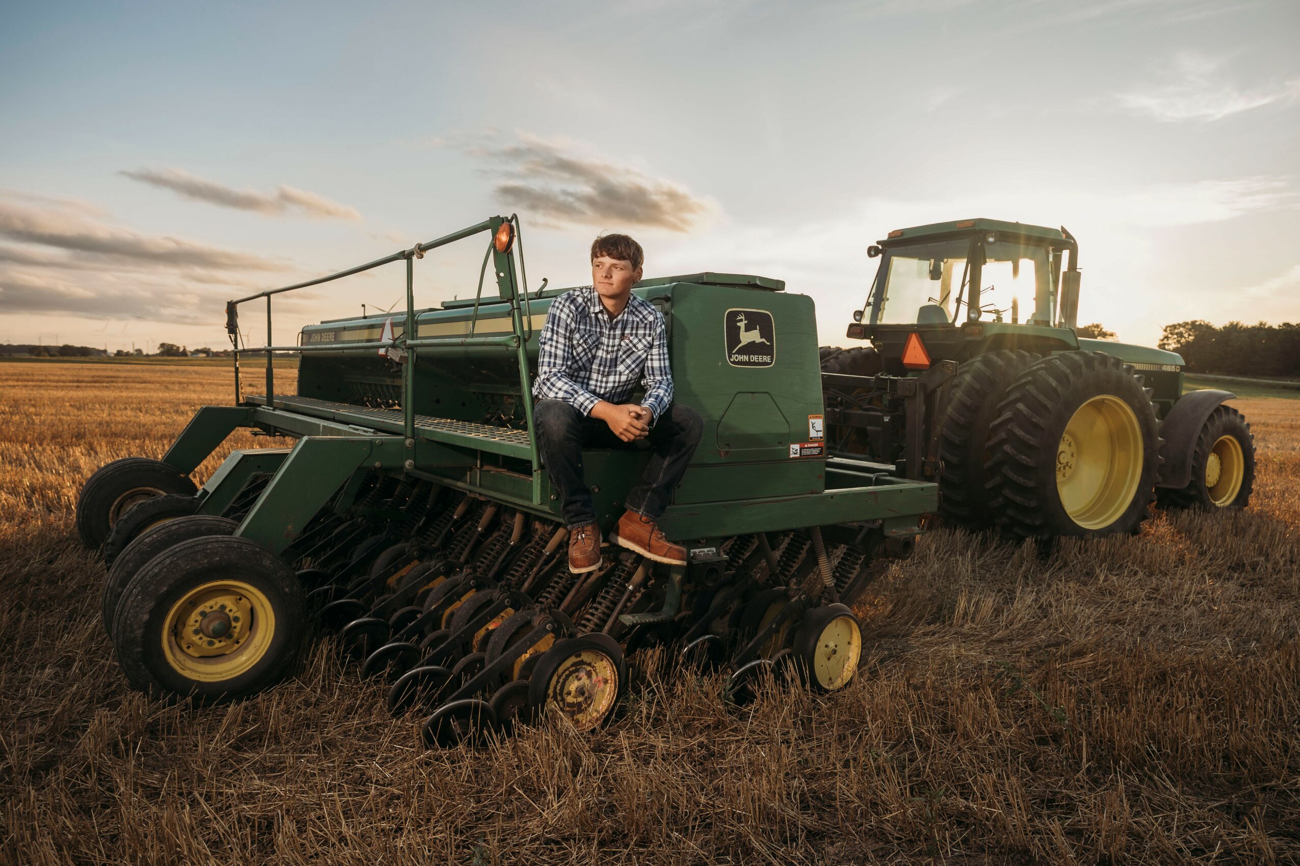 Person sitting on a green farming machine in a field with a tractor nearby during sunset.