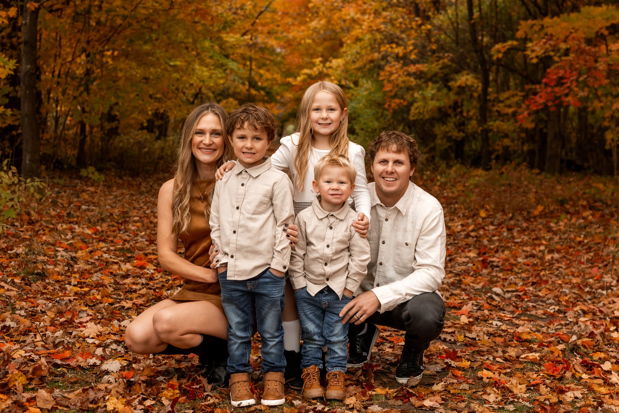 Group of five people, two adults and three children, standing outdoors on autumn leaves, smiling, with trees in fall colors behind them.