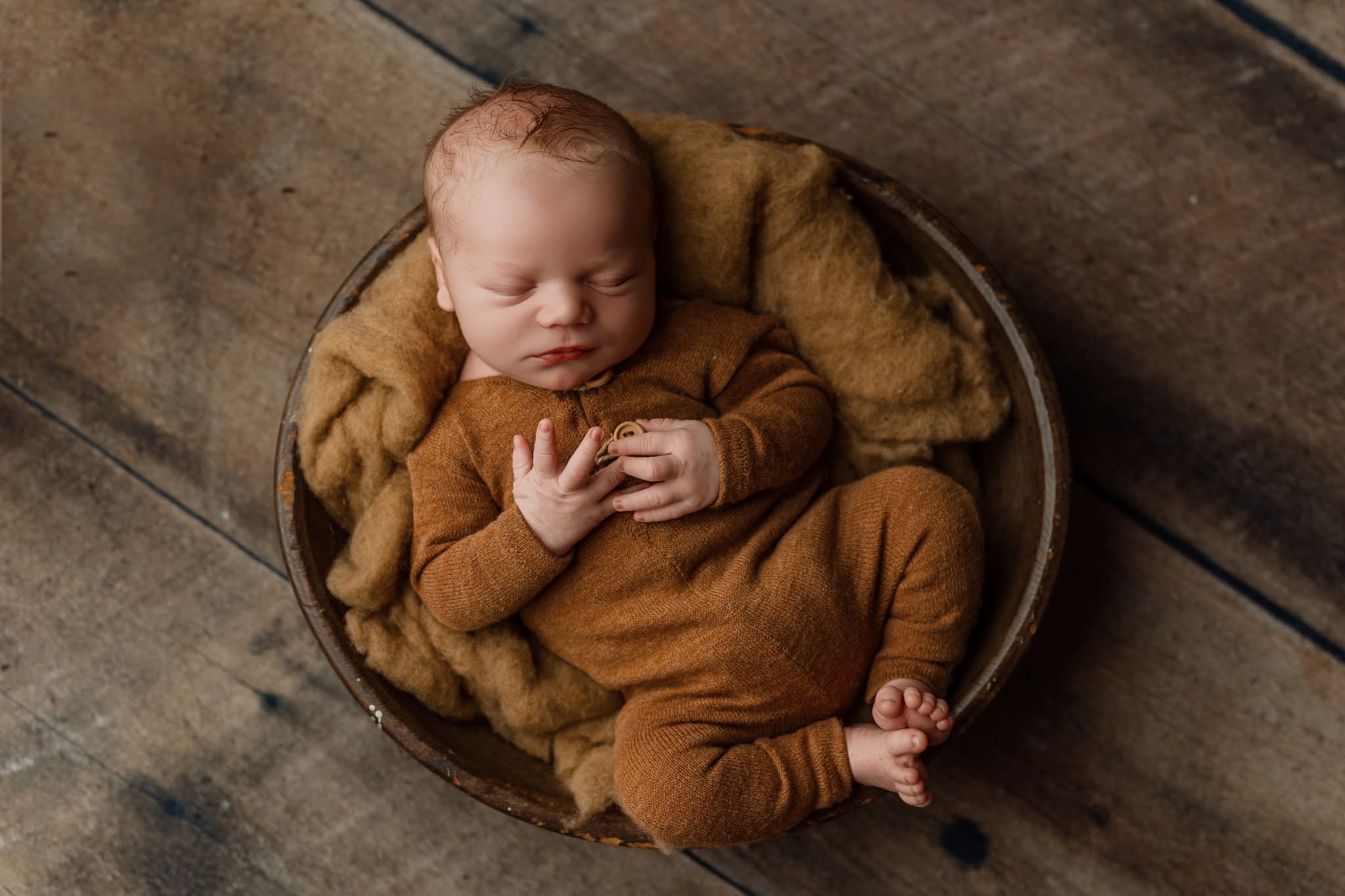 Baby sleeping in a round basket on a wooden floor, wrapped in a brown blanket, with hands near chest.