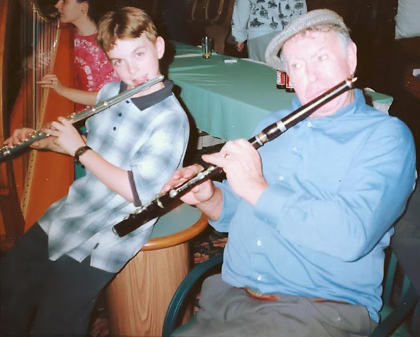 Two people playing flutes at a table in a social setting, with a woman in the background. One is a young boy, the other an older man.