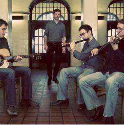 Four men playing musical instruments in a room with large arched windows, two sitting and two standing, with a tiled floor.