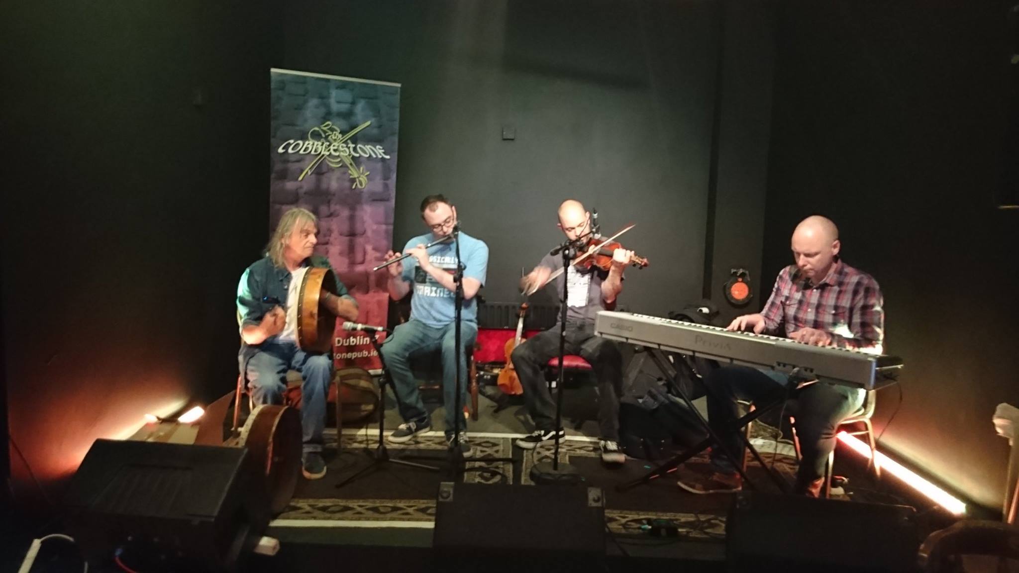 Four musicians perform on stage with instruments, including guitar, violin, and keyboard, in a dimly lit setting.