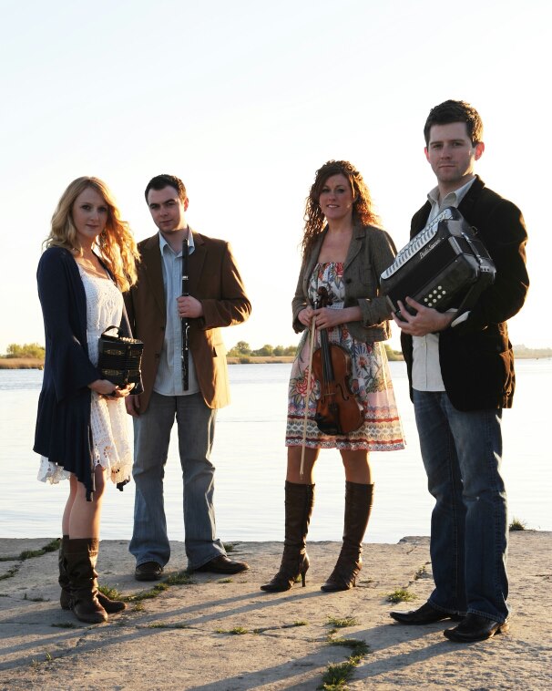 Four people stand outdoors near water, facing forward, with a clear sky background.