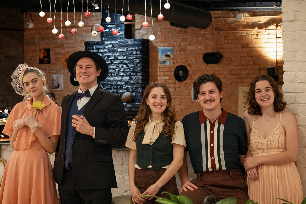Group of five people smiling at a party, standing indoors with brick walls and hanging lights.