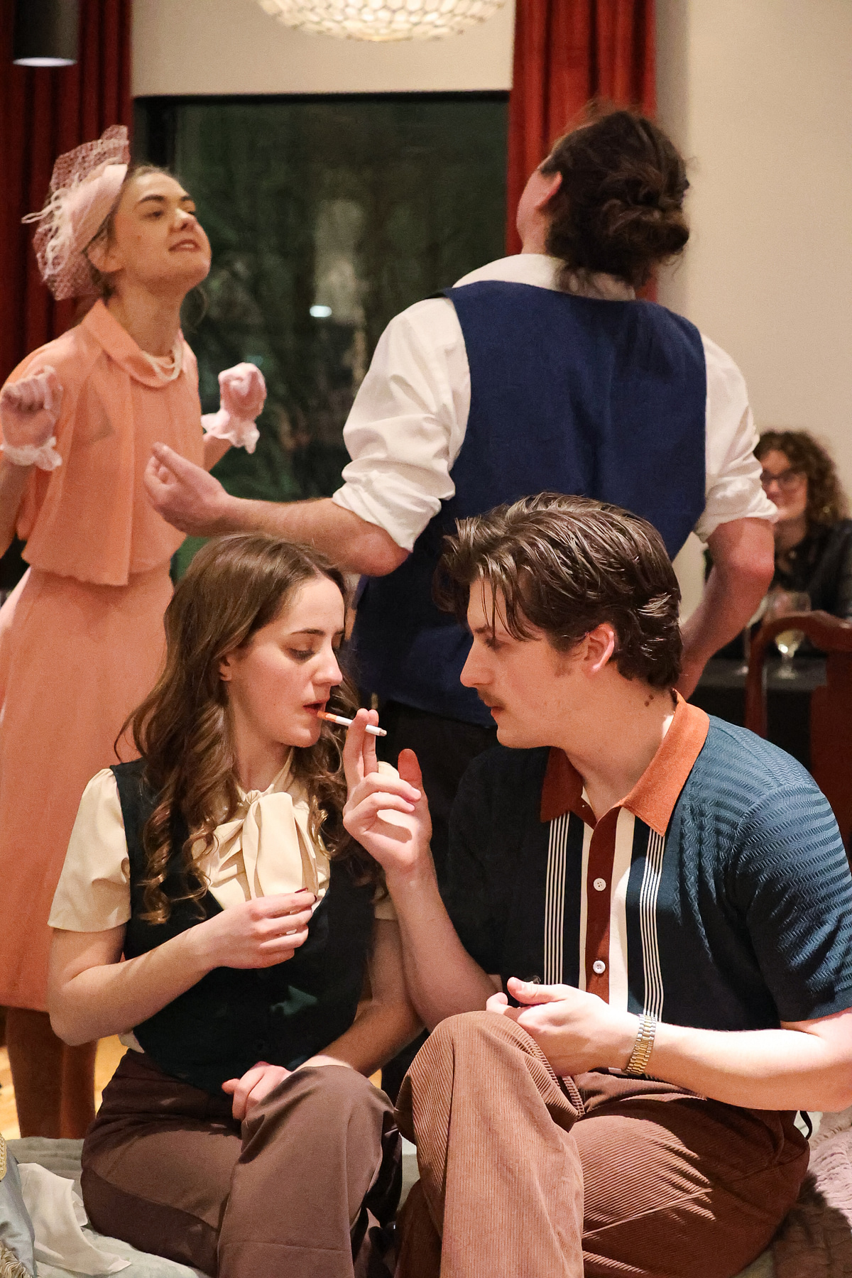 Two people sit close, one holding a cigarette, while a woman in a pink dress stands nearby with eyes closed.