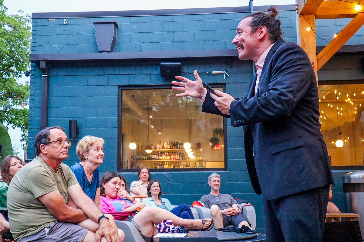 Man in suit speaking to seated group outdoors at night, with a blue building and string lights in background.