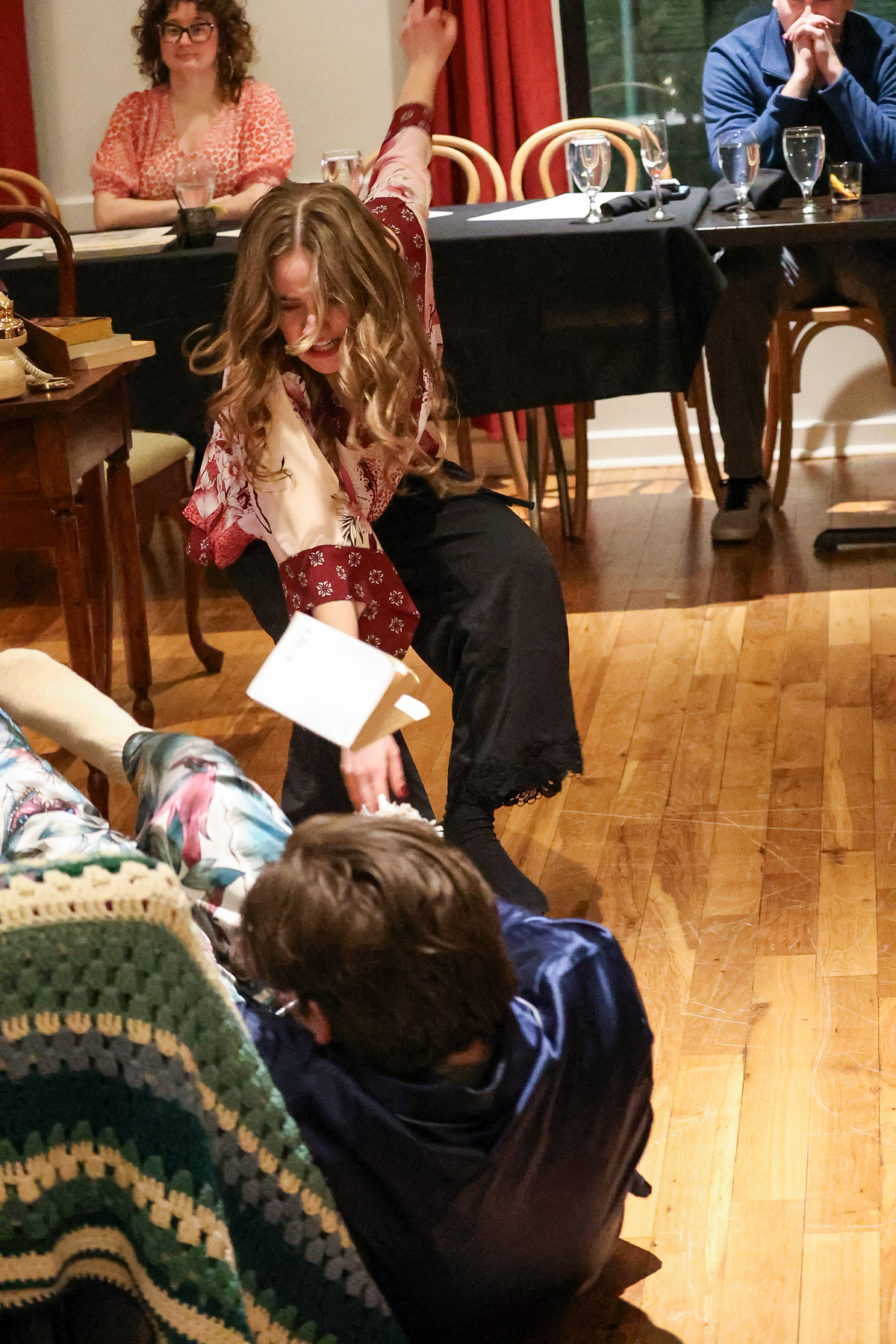 Child with long hair and patterned clothing sits on the floor, holding a white paper, with people seated at tables in background.