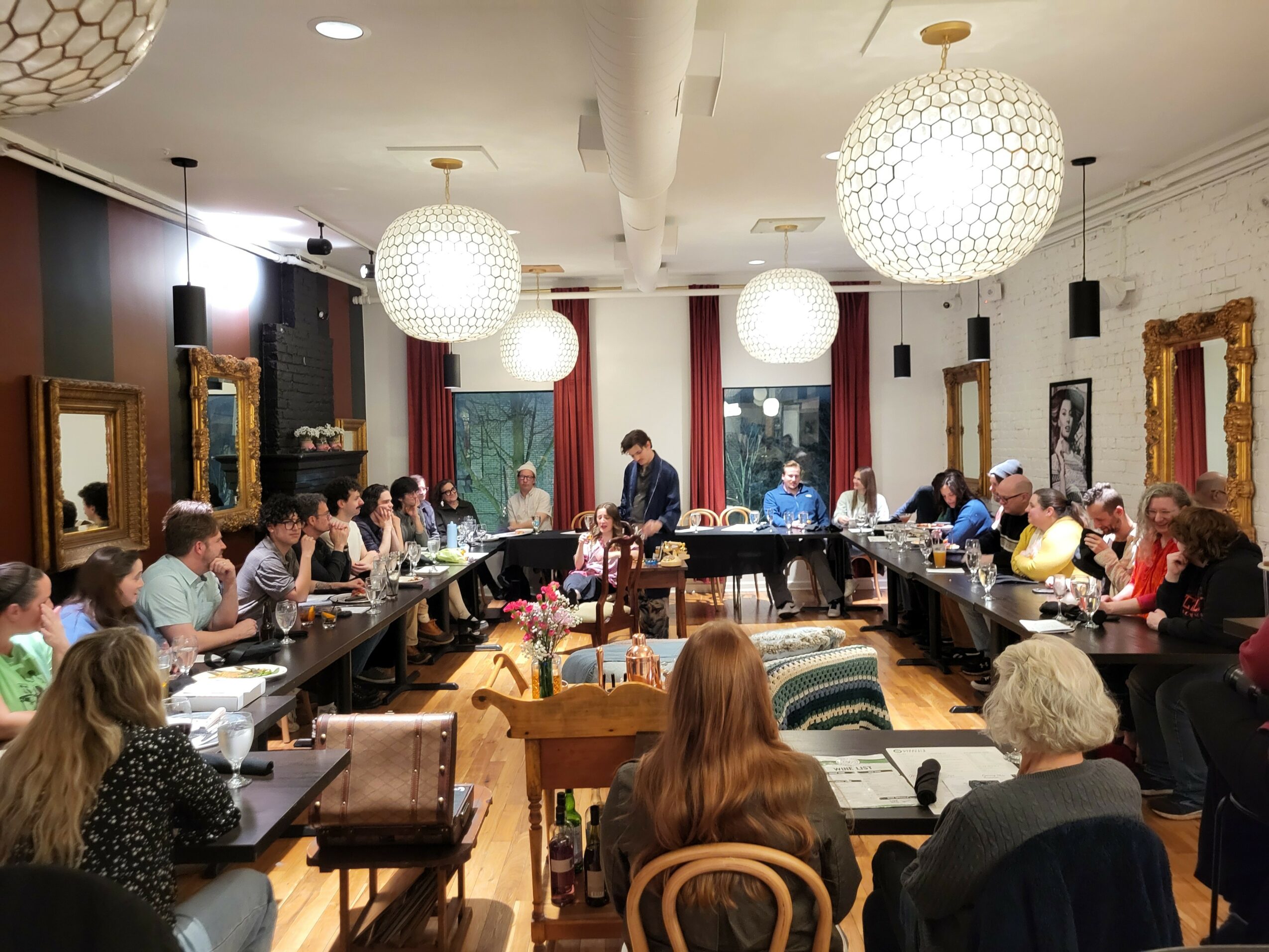 People seated around a long table in a decorated room with framed pictures, chandeliers, and curtains.
