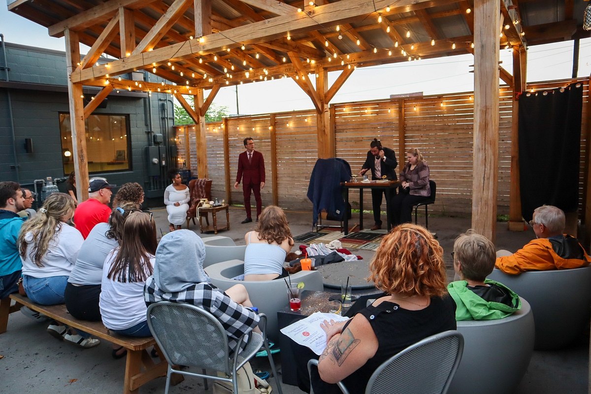 People seated outdoors watching a performance under a wooden pergola with string lights, with two performers at a piano and microphone.