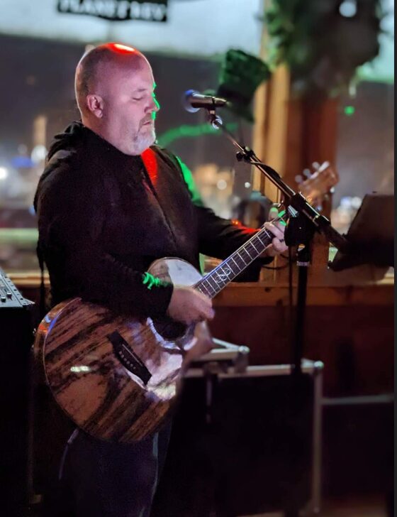 Man playing an acoustic guitar and singing into a microphone in a dimly lit indoor setting.