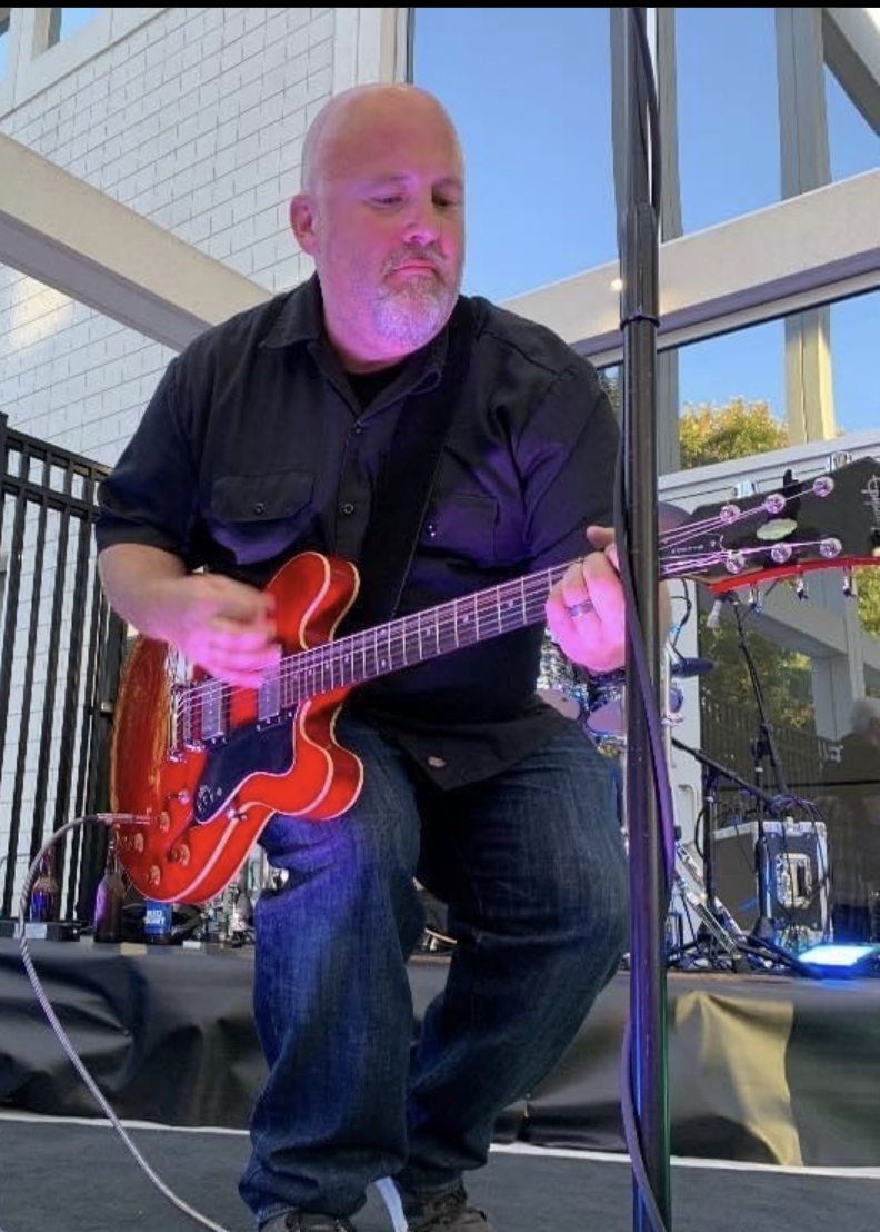 Man playing red electric guitar on stage outdoors, with glass building and trees in background.