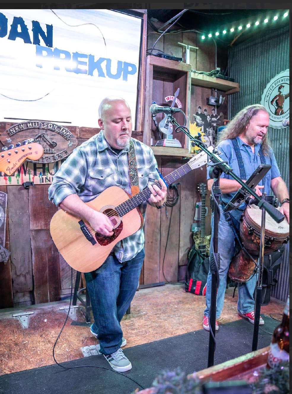 Two men playing guitars on stage with microphones, in a cozy venue with wooden decor and musical equipment.