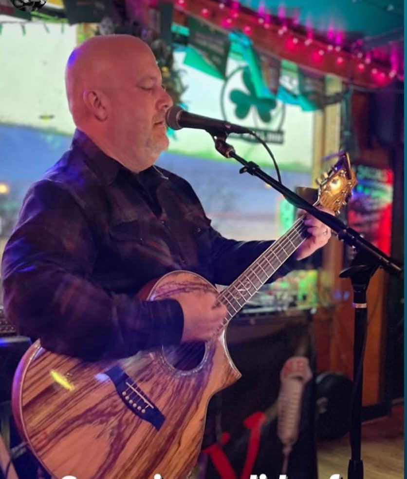 Bald man playing an acoustic guitar and singing into a microphone in a colorful, decorated venue.