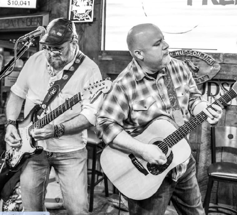 Two men playing guitars on stage, one with a cap and glasses, the other bald, in a casual setting.
