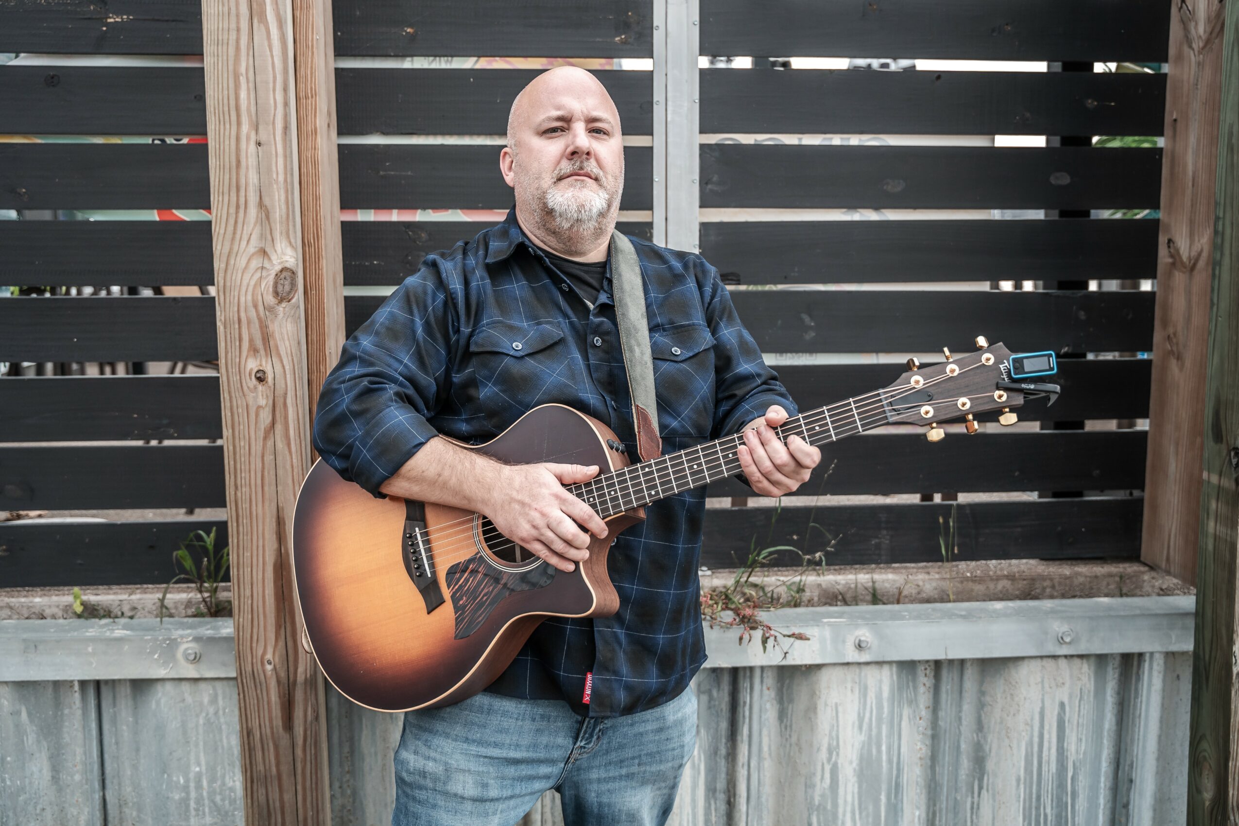 Man with a beard playing an acoustic guitar outdoors in front of a wooden fence.