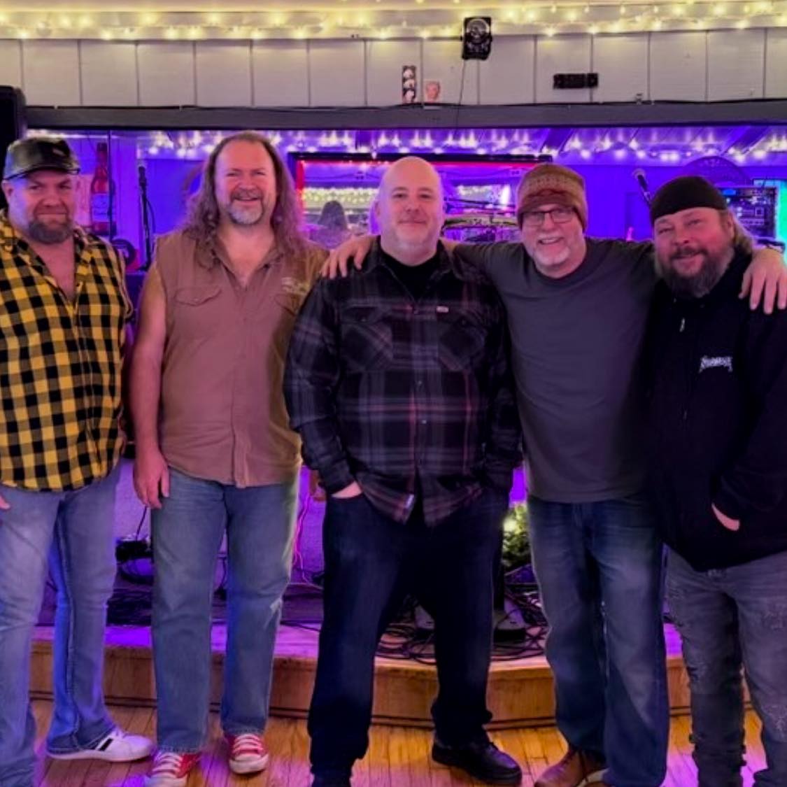 Five men standing together in a bar with purple lighting, smiling, arms around each other, in casual clothing.