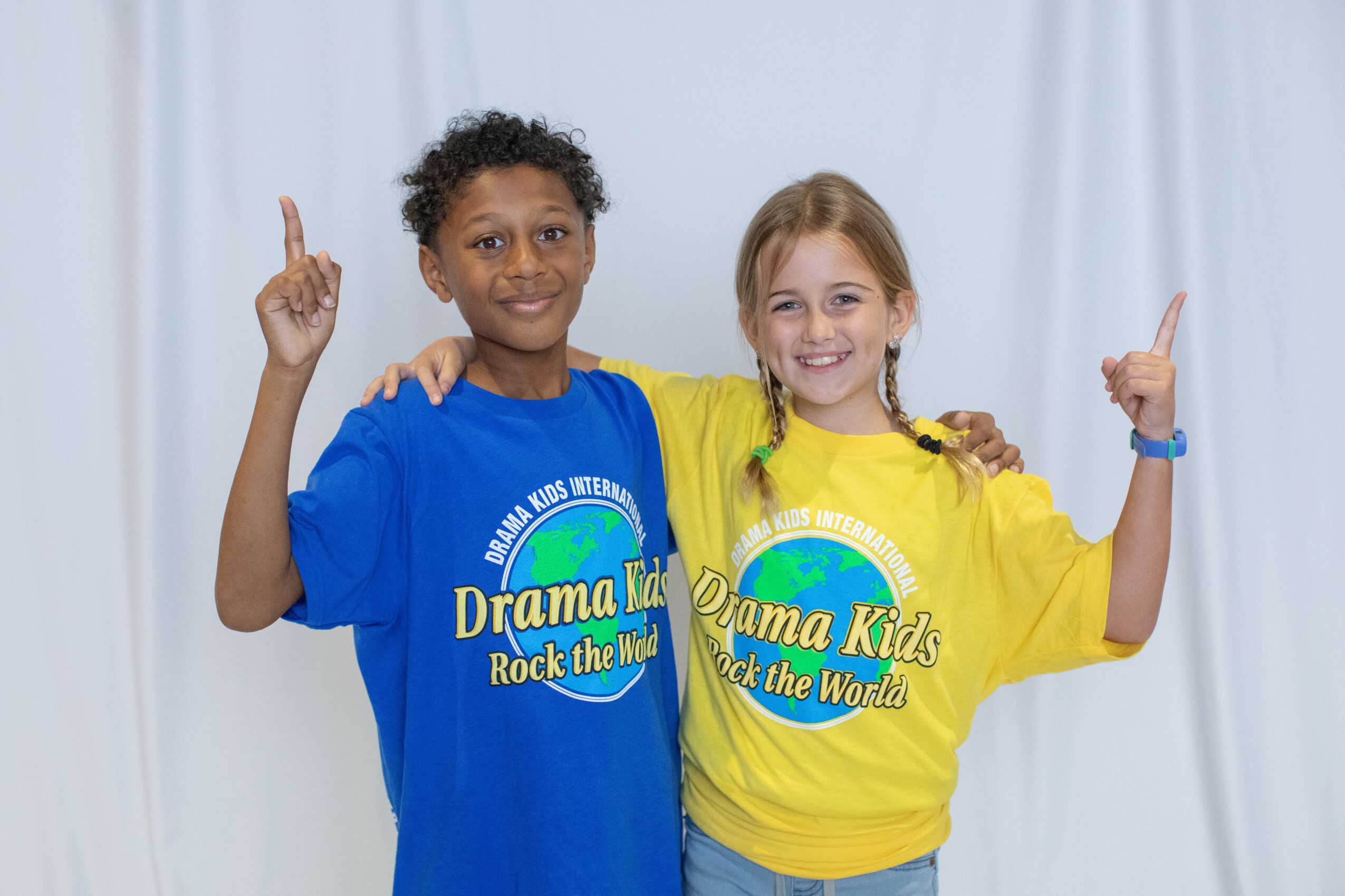 Two children, a boy and a girl, smiling and raising one finger each, standing against a white curtain background.