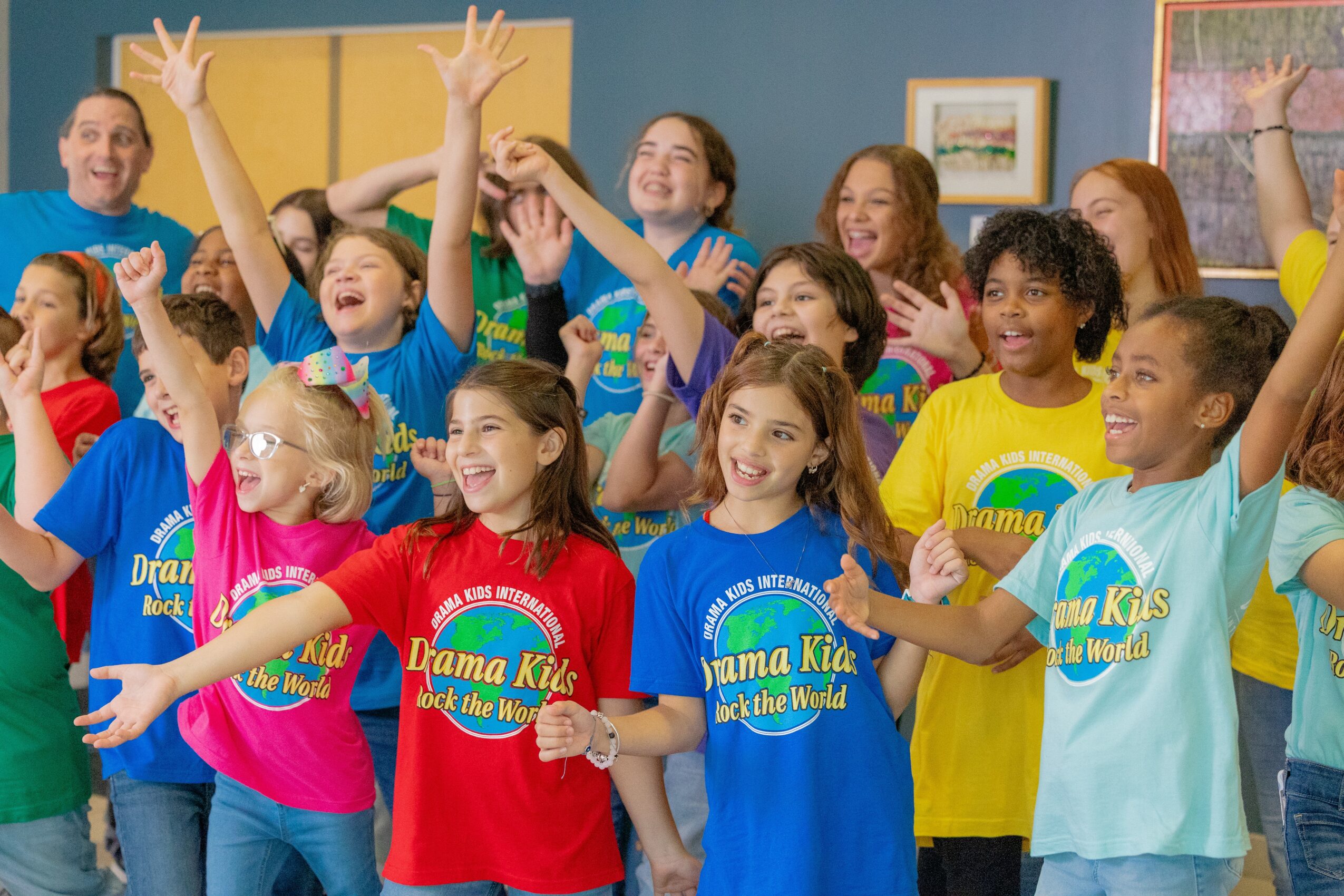 Group of children and adults smiling and raising hands in a colorful room, wearing matching T-shirts with 'Drama Kids' logo.