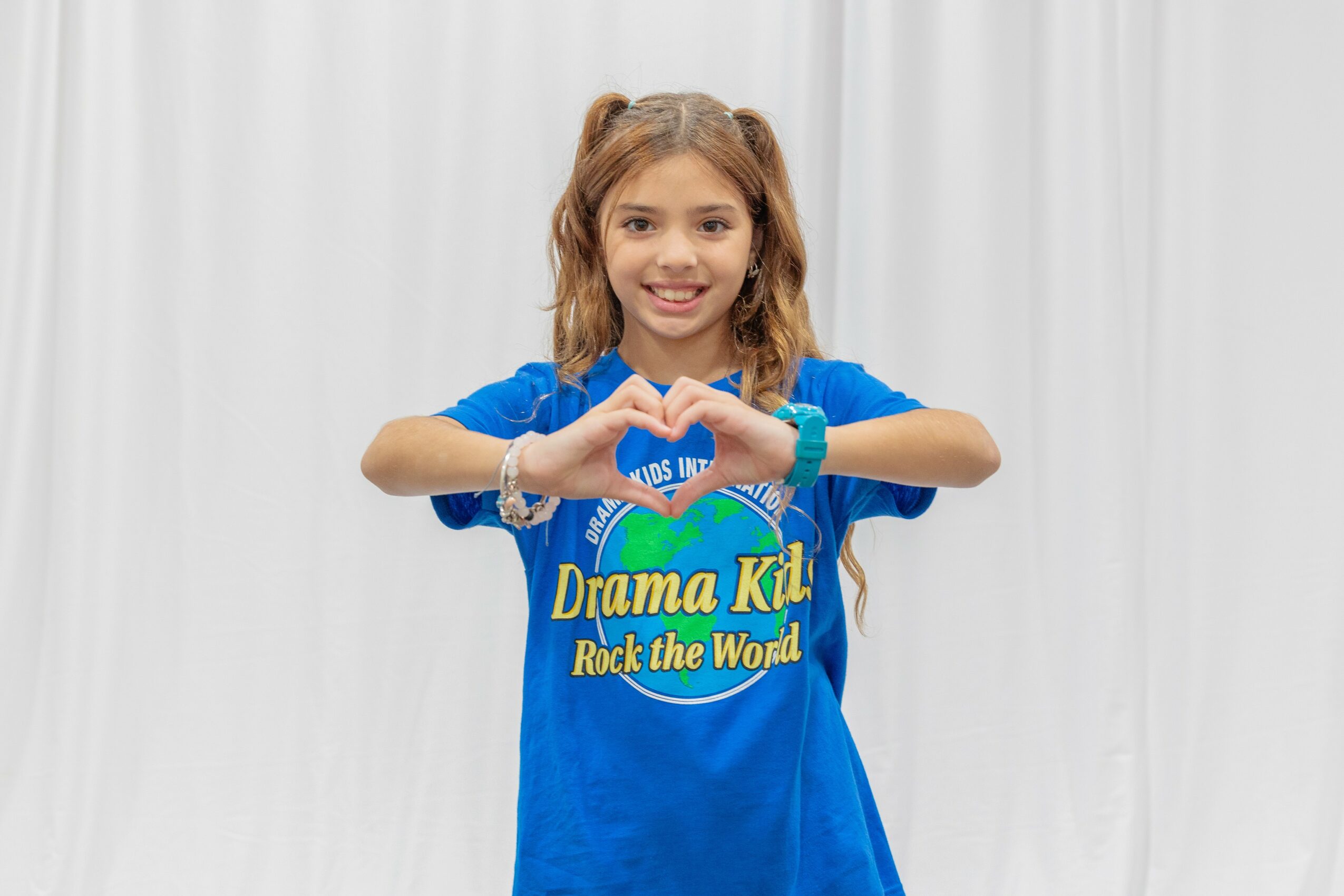 Young girl with brown hair in pigtails smiling and making a heart shape with her hands, wearing a blue T-shirt, standing in front of a white curtain.