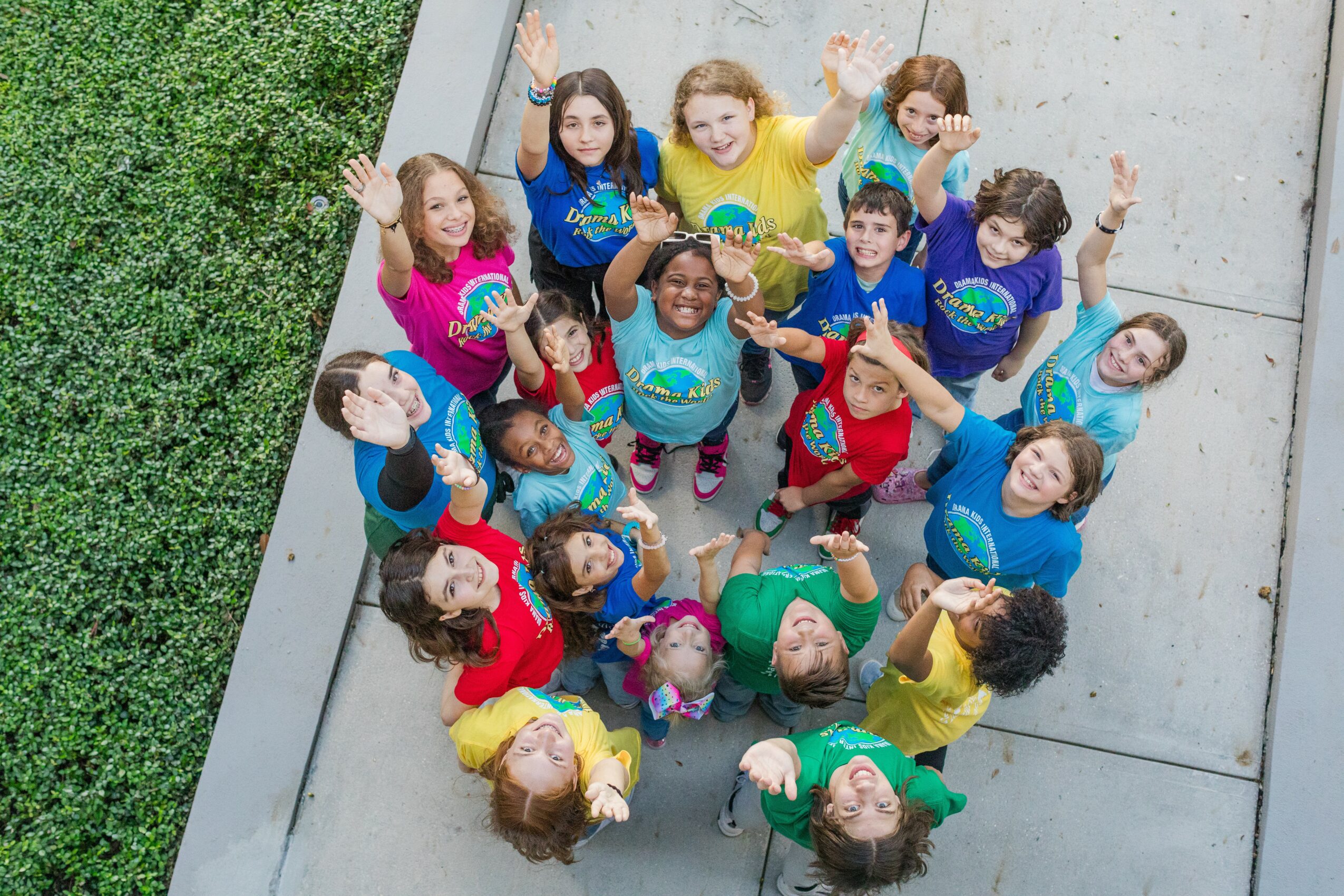 Group of children looking up, smiling, and waving outdoors on concrete and grass.