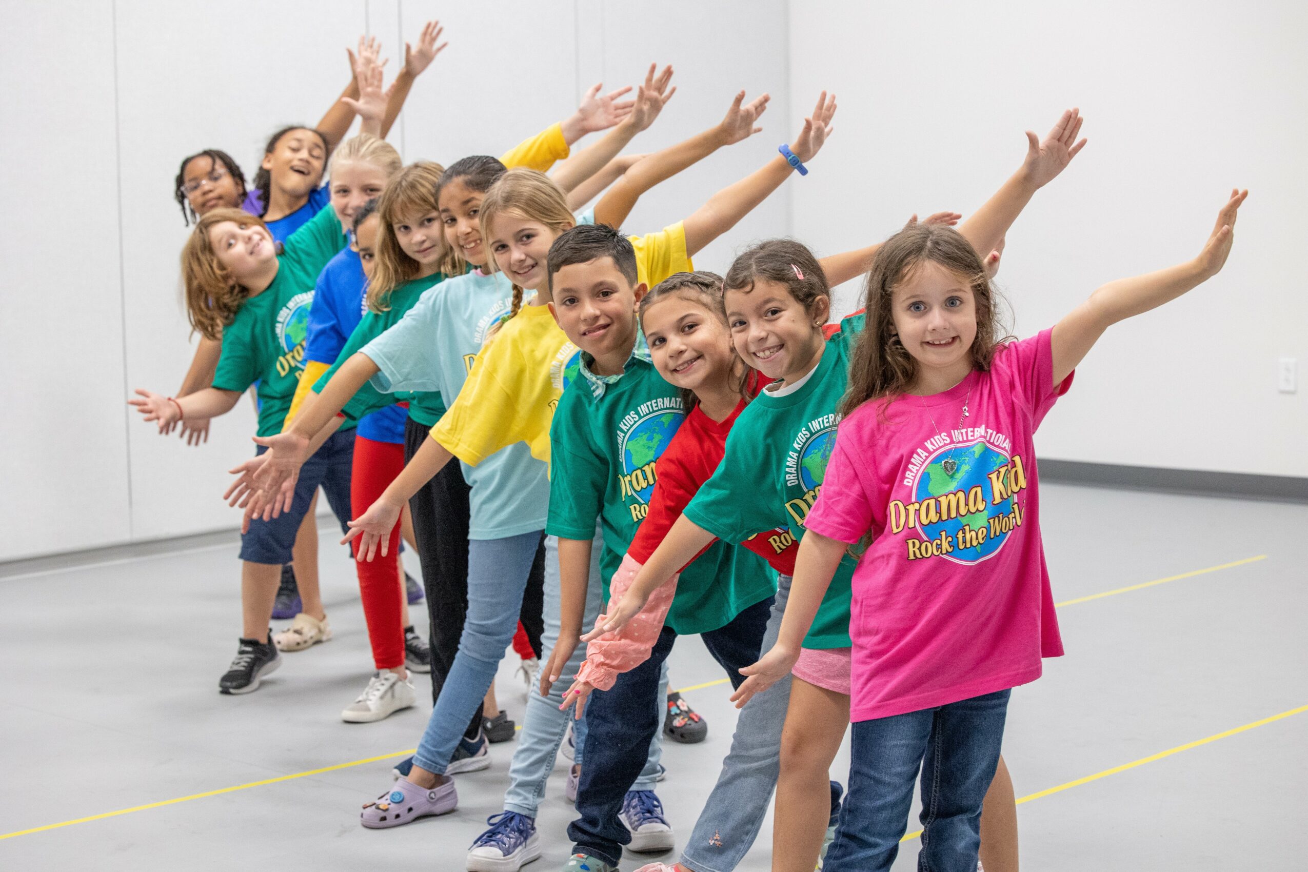 Group of children in colorful shirts standing in line with arms raised, smiling, in a room with white walls.