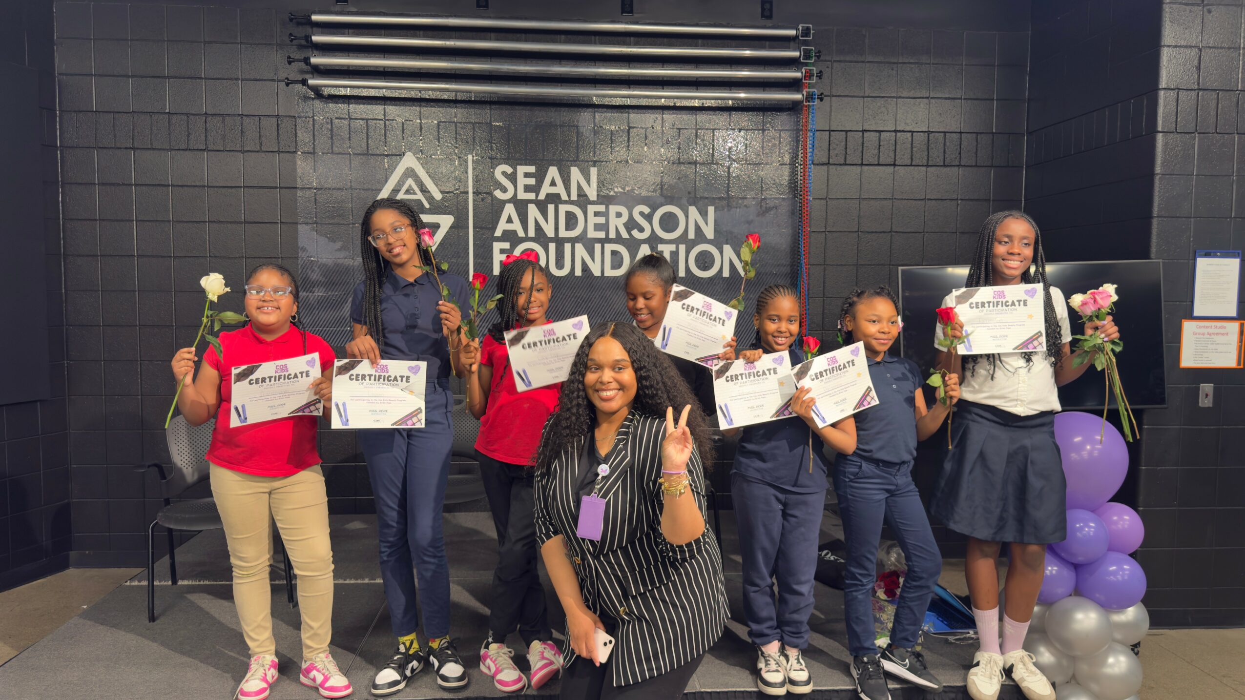 Group of seven children and one adult woman holding certificates and flowers, standing in front of a black wall with a logo and text.