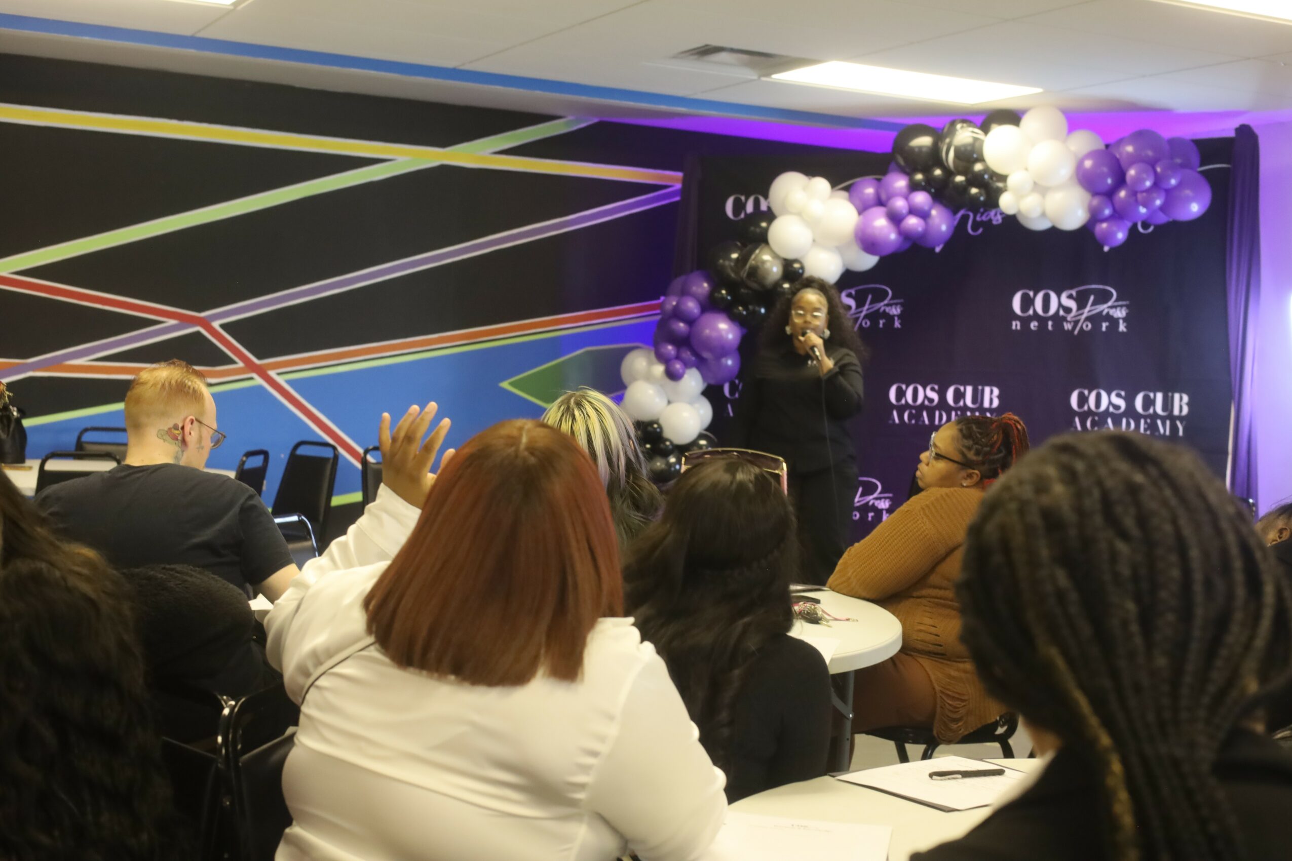 Person speaking on stage with purple and white balloon decorations, audience seated and listening attentively.