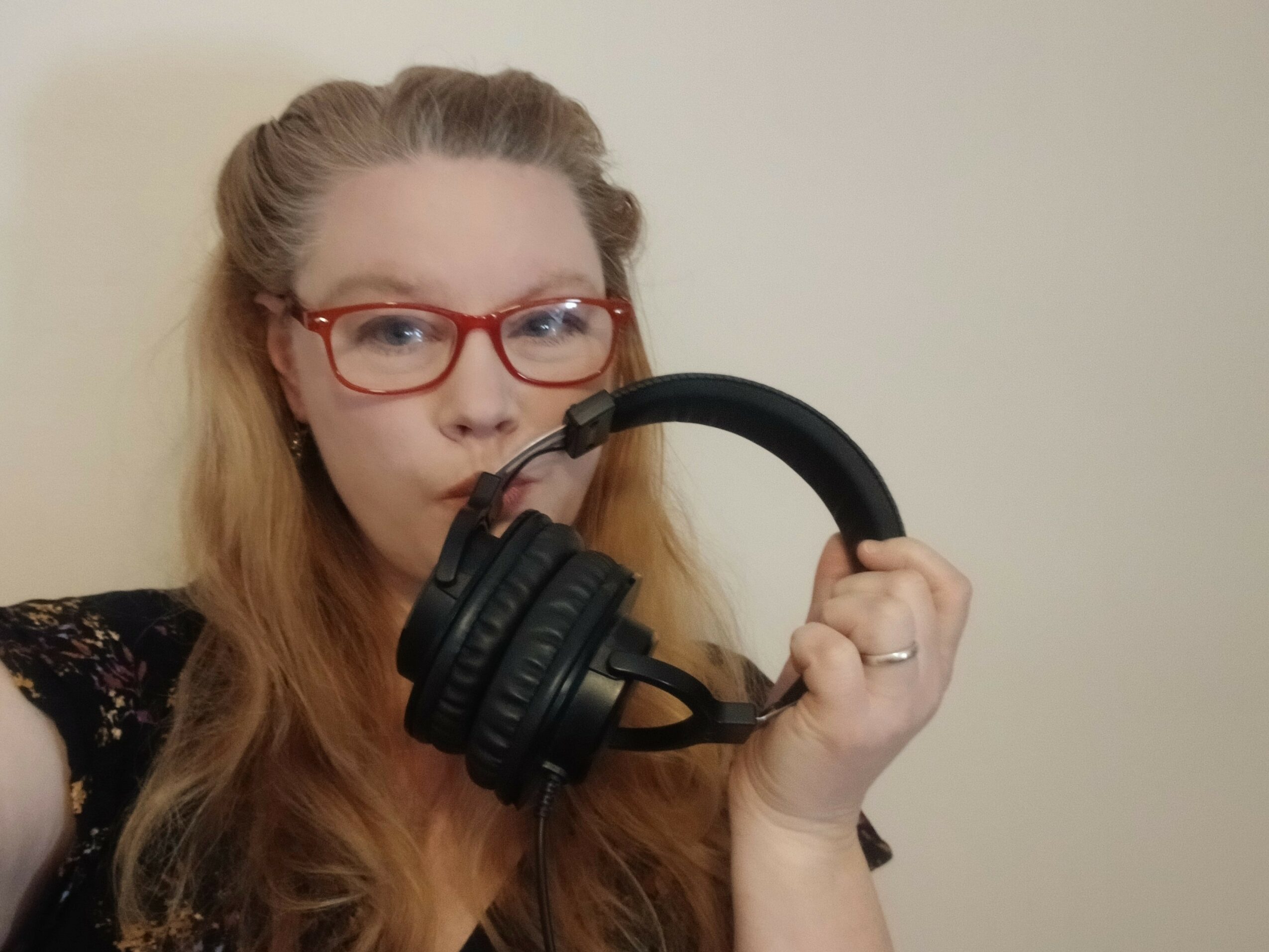Woman with long hair and red glasses holding headphones near her face, against a plain background.
