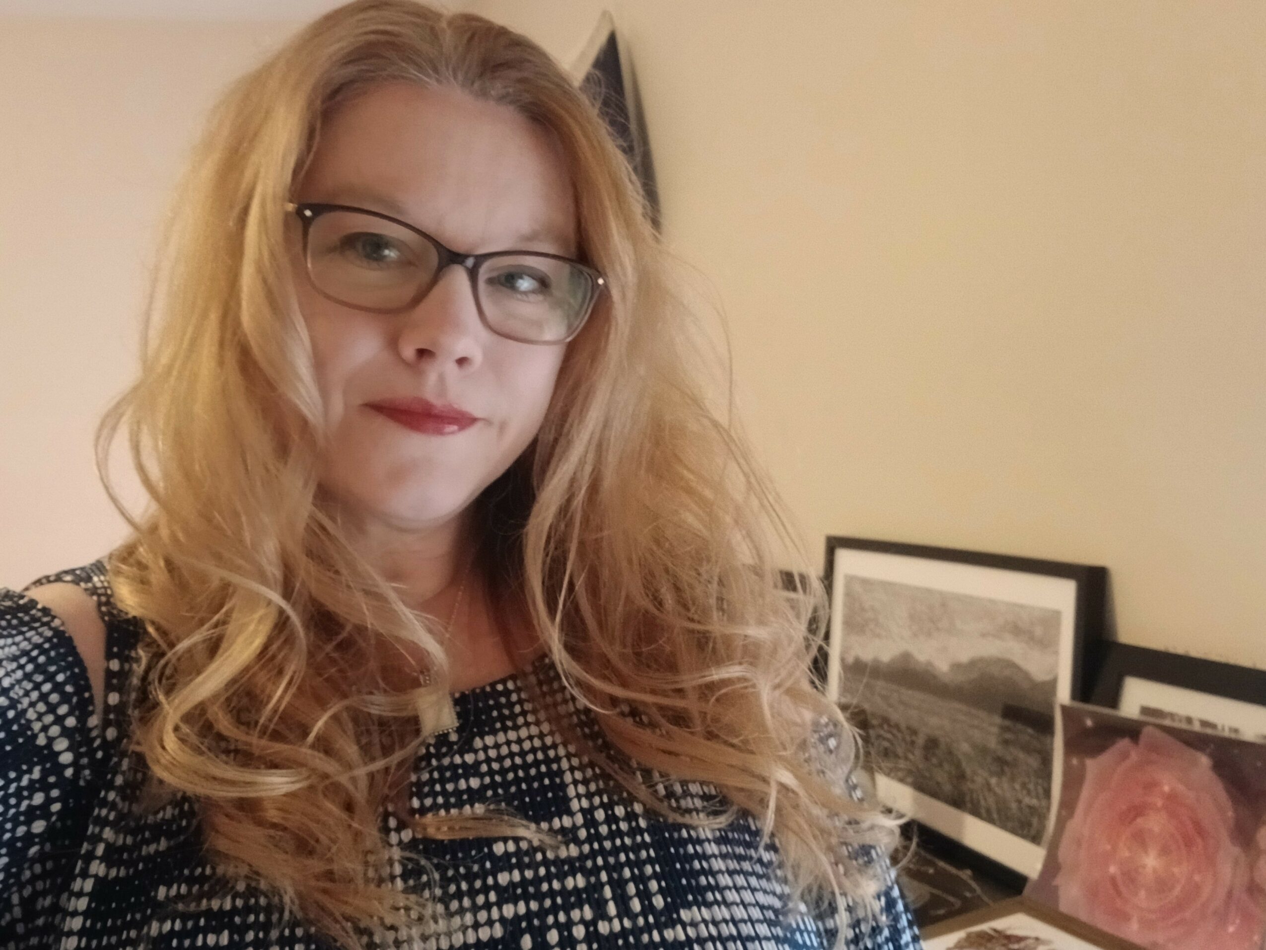 Woman with glasses and wavy hair looking to the side, in front of framed pictures on a wall.