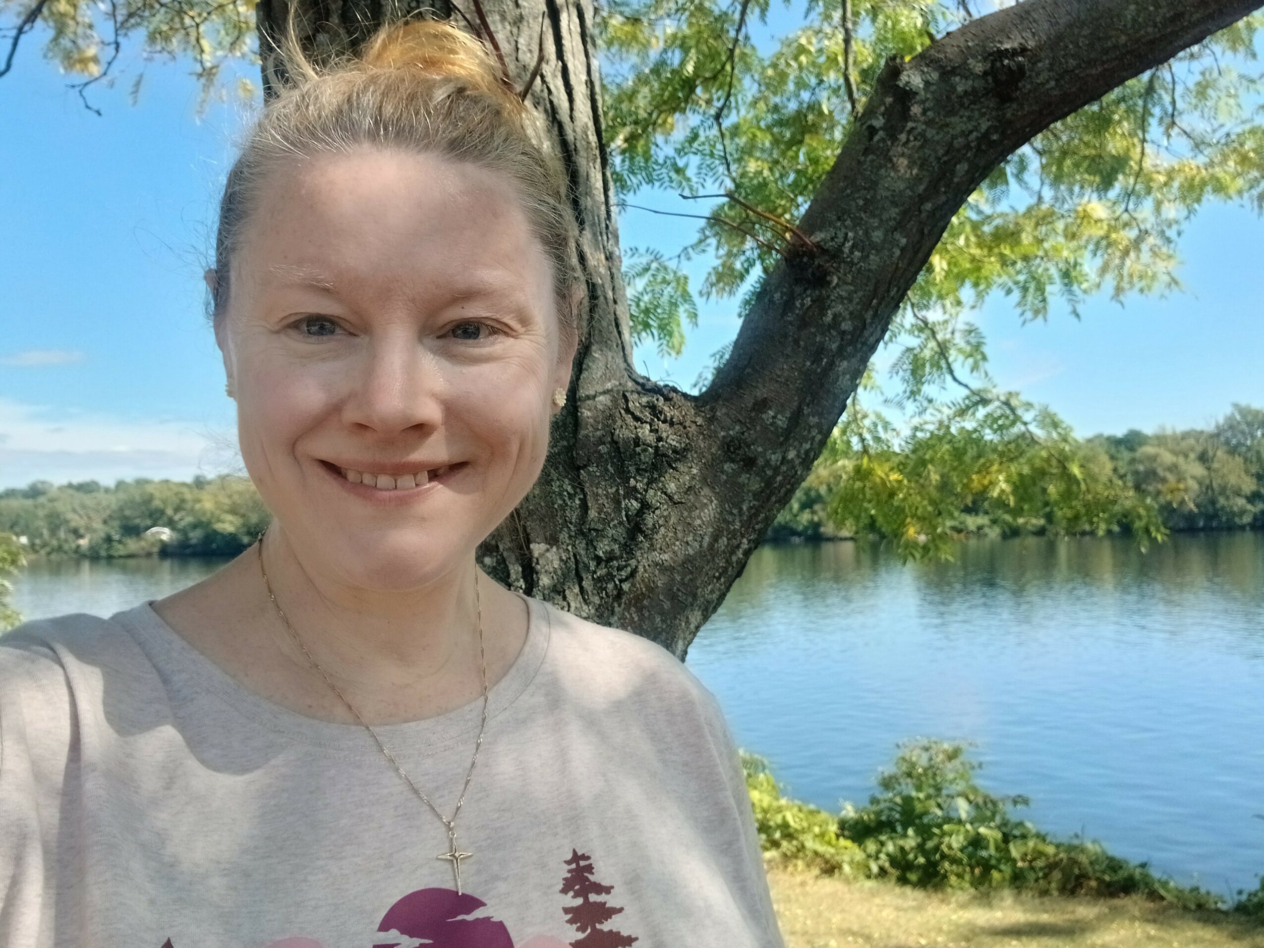 Young woman smiling outdoors near a lake with trees and blue sky in background.