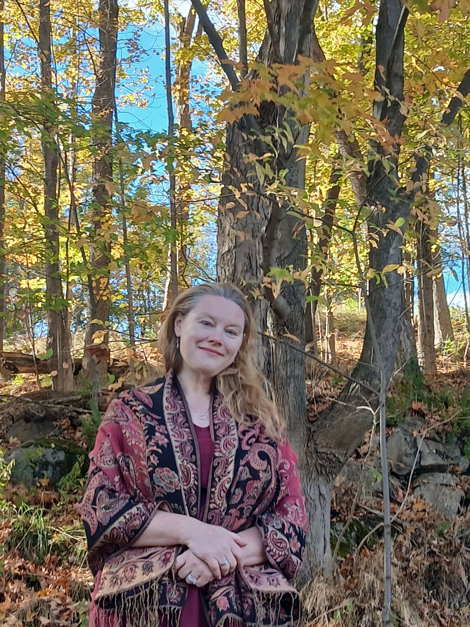 Woman with long hair in a patterned jacket standing among trees with autumn leaves.