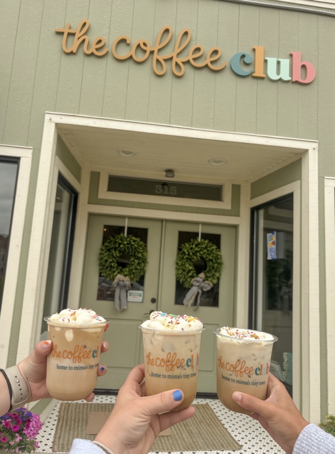 Three people holding cups of coffee outside a cafe with a sign reading 'the coffee club' in colorful letters, green building exterior.